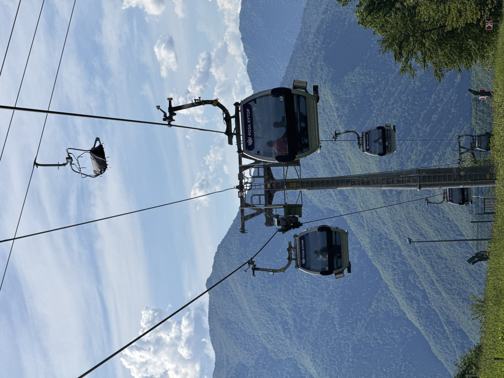 An outdoor photograph captures a gondola lift system in operation, set against a backdrop of forested mountains under a bright, partly cloudy sky. The image focuses on a modern gondola cabin, visible in the midground, with large windows reflecting the sunny surroundings. Silhouettes of passengers are discernable inside. The cabin prominently displays the text "РОЗА ХУТОР" (Roza Khutor) in white Cyrillic letters, accompanied by a logo, and smaller numbers such as "G9" and "G2" are also visible.
A sturdy support tower for the lift system stands near the main gondola cabin. Steel cables extend across the frame, supporting multiple cabins and a lone open chairlift in the upper left section of the image. In the background, vast, green-covered mountains rise under a clear blue sky dotted with scattered white clouds. Patches of green grass and trees are visible in the foreground, suggesting an elevated viewpoint within a natural setting. The scene depicts a daytime setting with good weather, indicating a leisure or transport activity within a mountainous resort area, consistent with Roza Khutor in Estosadok, Russia.