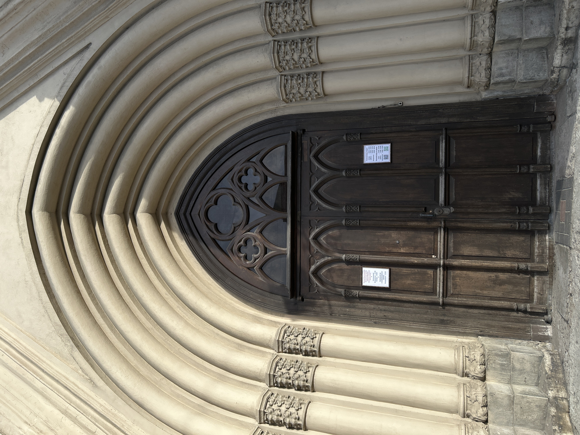 A gothic-style arched wooden door in Saint Petersburg, Russia. The door is framed by multiple layers of arched stonework, each layer decorated with floral carvings. The wooden door is divided into two sections, the upper section featuring a trefoil design. Two small white signs with black text are affixed to the right side of the door. The door is closed and the overall scene appears to be a close-up shot of an architectural detail.