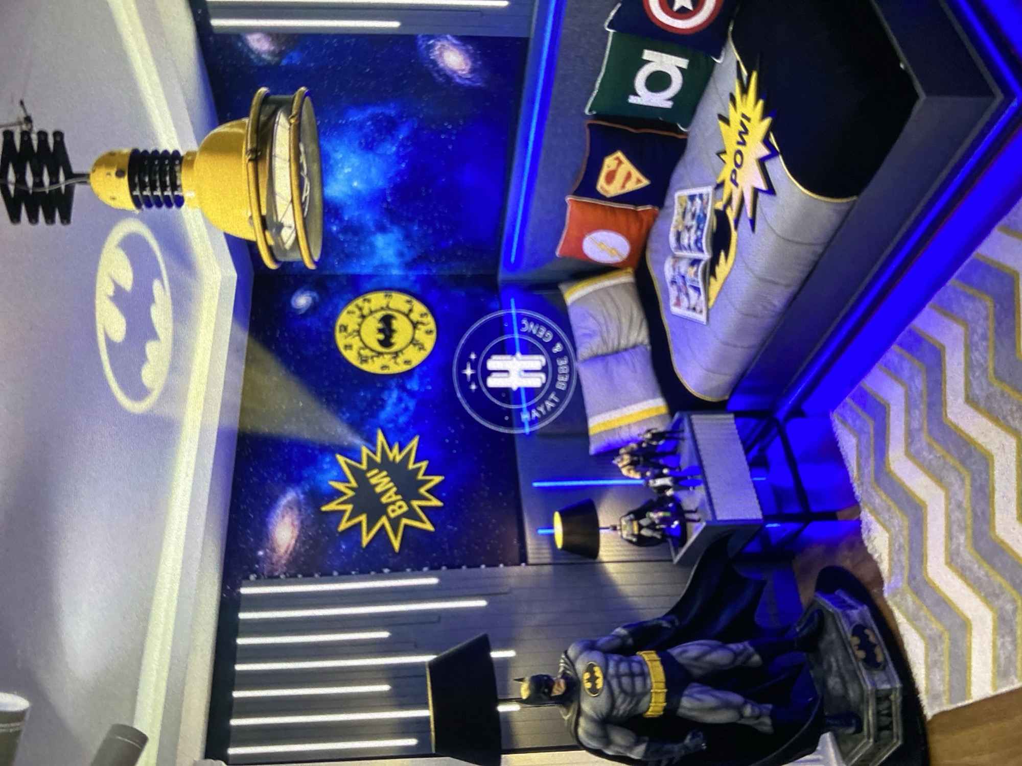 A room with superhero-themed decor. A large Batman figurine stands on a platform. A couch is adorned with superhero-themed pillows. The walls feature Batman logos and superhero-style action words. The location is Lhokseumawe, Indonesia.