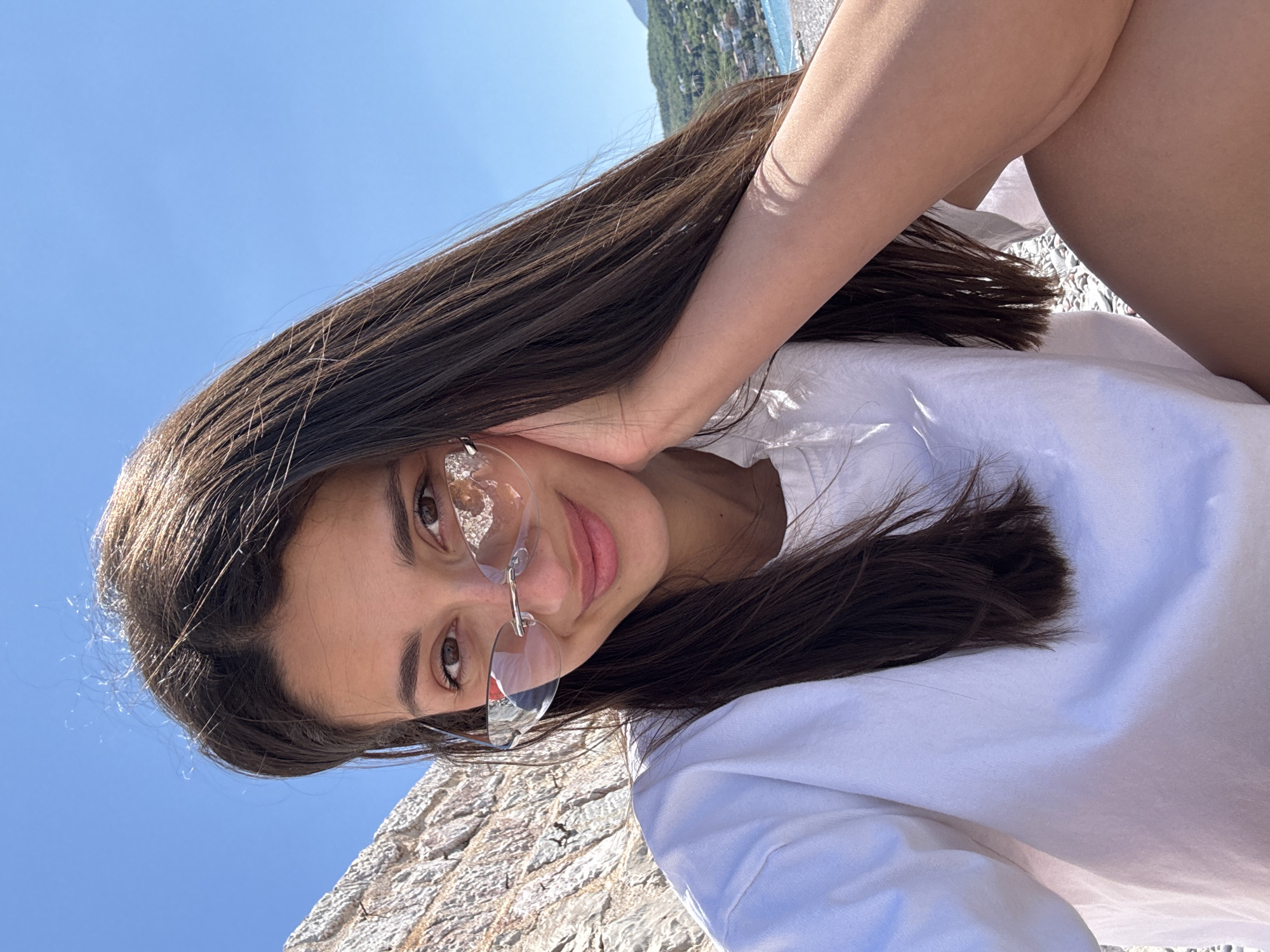A young woman with long, dark brown hair is facing the camera while wearing frameless glasses and a white t-shirt. She is holding her hand to her face. The image is taken at an angle. The background features a clear blue sky and a portion of a stone structure. In the distance, a blurred coastline and buildings suggest an urban area, consistent with the city of Sutomore, Montenegro.