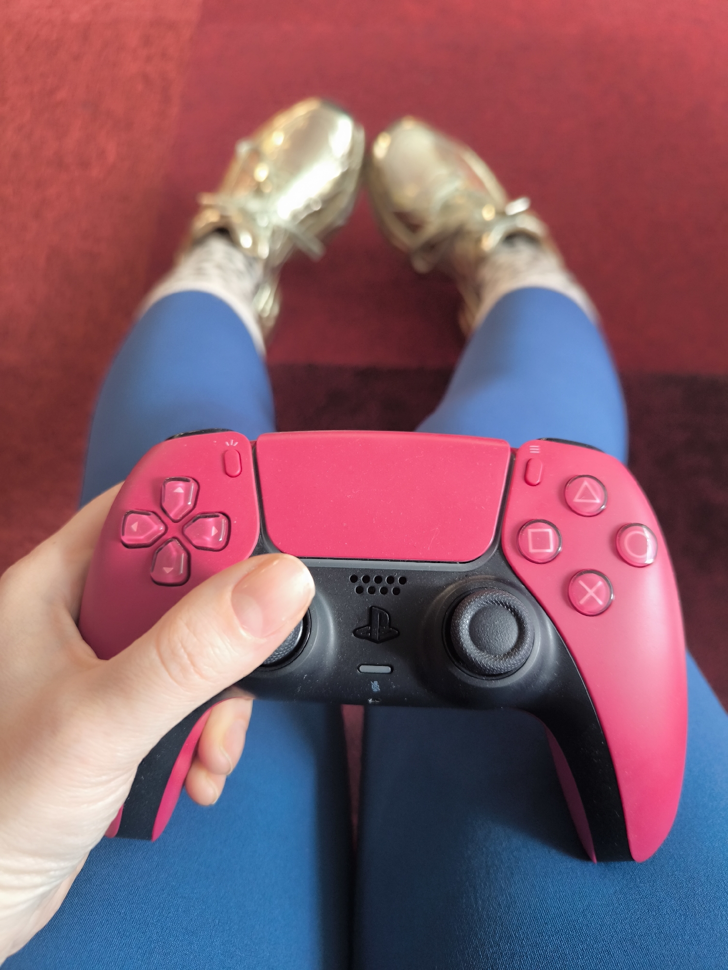 A person sits on a red and blue patterned surface, likely a blanket or rug, wearing blue leggings and shiny gold shoes. They are holding a pink and black PlayStation 5 DualSense controller. The scene is indoors, and the location within the city of Unknown, Unknown is indeterminable based on visual information.