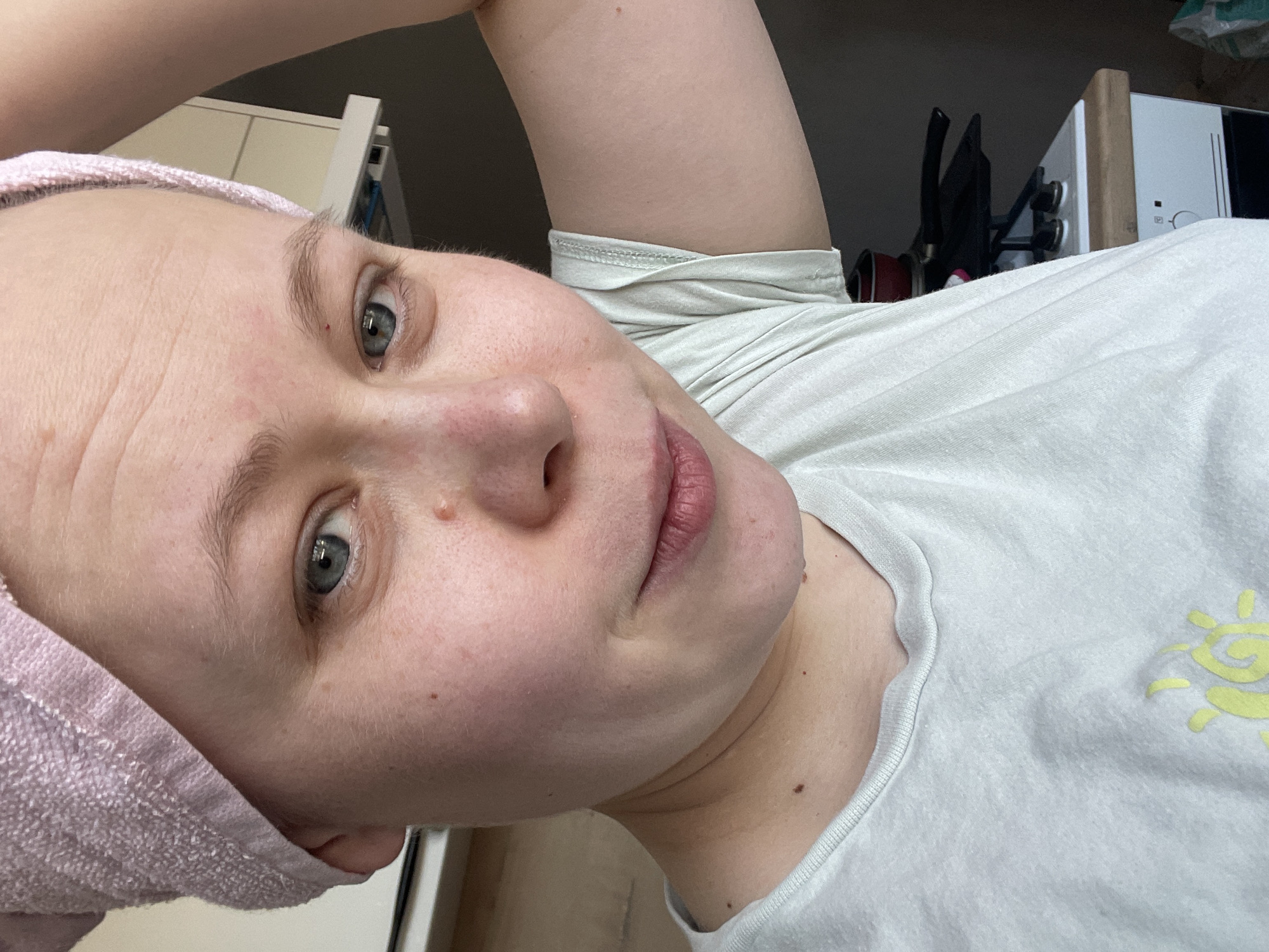 A selfie of a woman with fair skin and blue eyes. She is wearing a pink towel wrapped around her head and a light gray t-shirt with a yellow sun design. Her face is positioned at an angle to the camera, with the right side slightly closer. Behind her is a partially visible kitchen counter and appliances. The location is likely indoors and no information in the image suggests it is in the city of Chaikovsky, Russia.