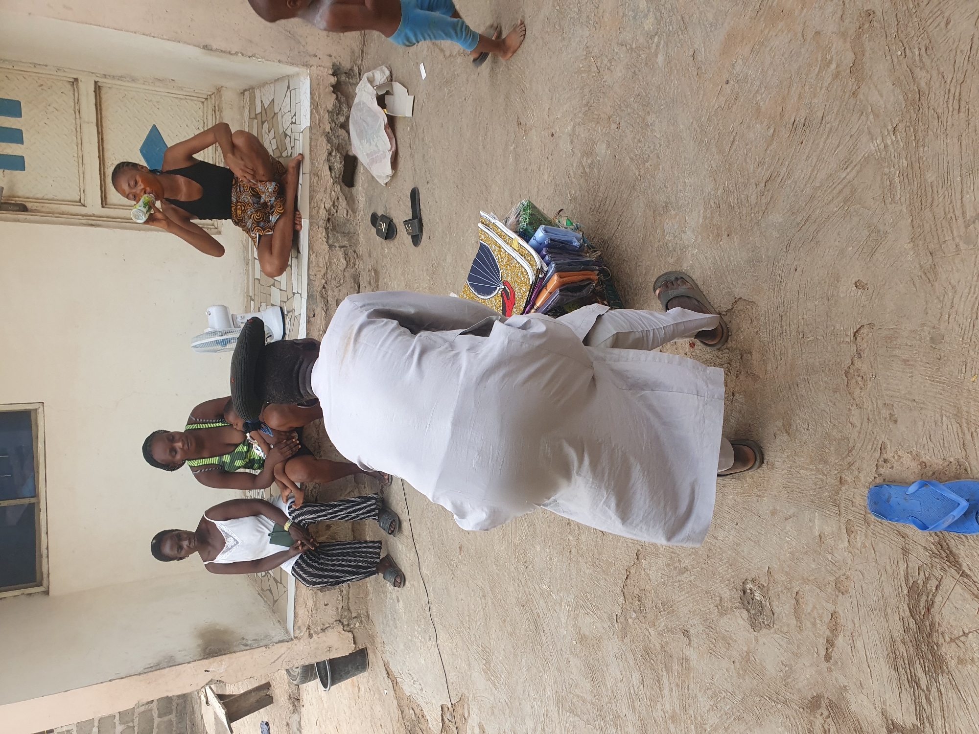 The image captures an outdoor scene, likely a courtyard or open-air space, showing several individuals gathered on a light-colored, uneven concrete ground. The setting appears to be a warm climate, possibly Lokoja, Nigeria, given the context.
In the foreground, with their back to the viewer, stands a person wearing a long, light-colored garment and dark sandals. To their right on the ground, a stack of colorful, folded fabrics or clothing items, some with distinctive patterns like yellow with black and red designs, is neatly piled. Near these items are a white plastic bag, a brown cardboard box, and a pair of black slides with "KL" visible on one. A single blue flip-flop is further to the right.
To the left, two women are seated on the ground. The woman closer to the camera wears a white top and black-and-white striped pants, looking towards the viewer. Behind her, another woman in a green striped top holds a small child. Further to the left, on a raised concrete step against a building wall, a woman in a black tank top and patterned skirt is seated, drinking from a silver-colored can. A small white electric fan sits nearby. The building wall features a simple window and a closed door with blue accents. In the upper right background, a child wearing light blue pants is visible.
The scene depicts a casual gathering, possibly involving the display or sale of the fabrics. The bright daylight suggests it is daytime. The people appear relaxed, with no strong emotions discernible.