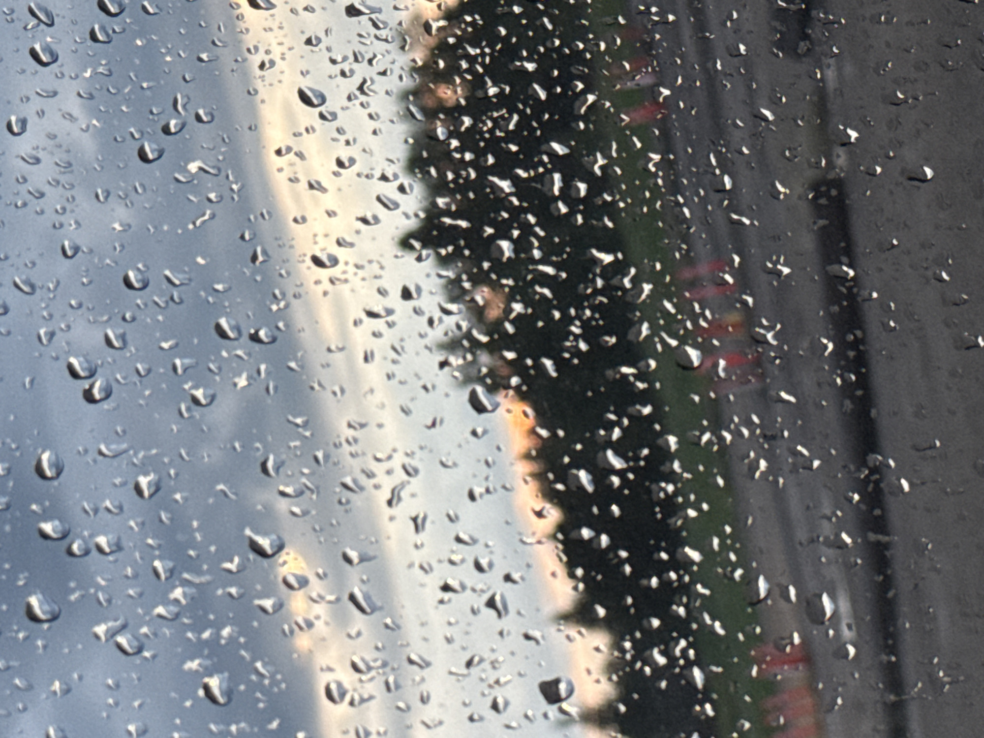 A close-up, rain-streaked view of a twilight sky with a hint of orange and pink sunset colors visible through the water droplets. In the mid-ground, a dark line of trees is silhouetted against the sky. The foreground shows a blurred asphalt surface with what appear to be orange traffic cones. The scene is likely from inside a vehicle or building looking out through a window. The location is identified as Stony Plain, Canada, implying an outdoor setting with a visible sky and trees characteristic of the region. No people or specific actions are discernible due to the focus on the rain and the blurred background.