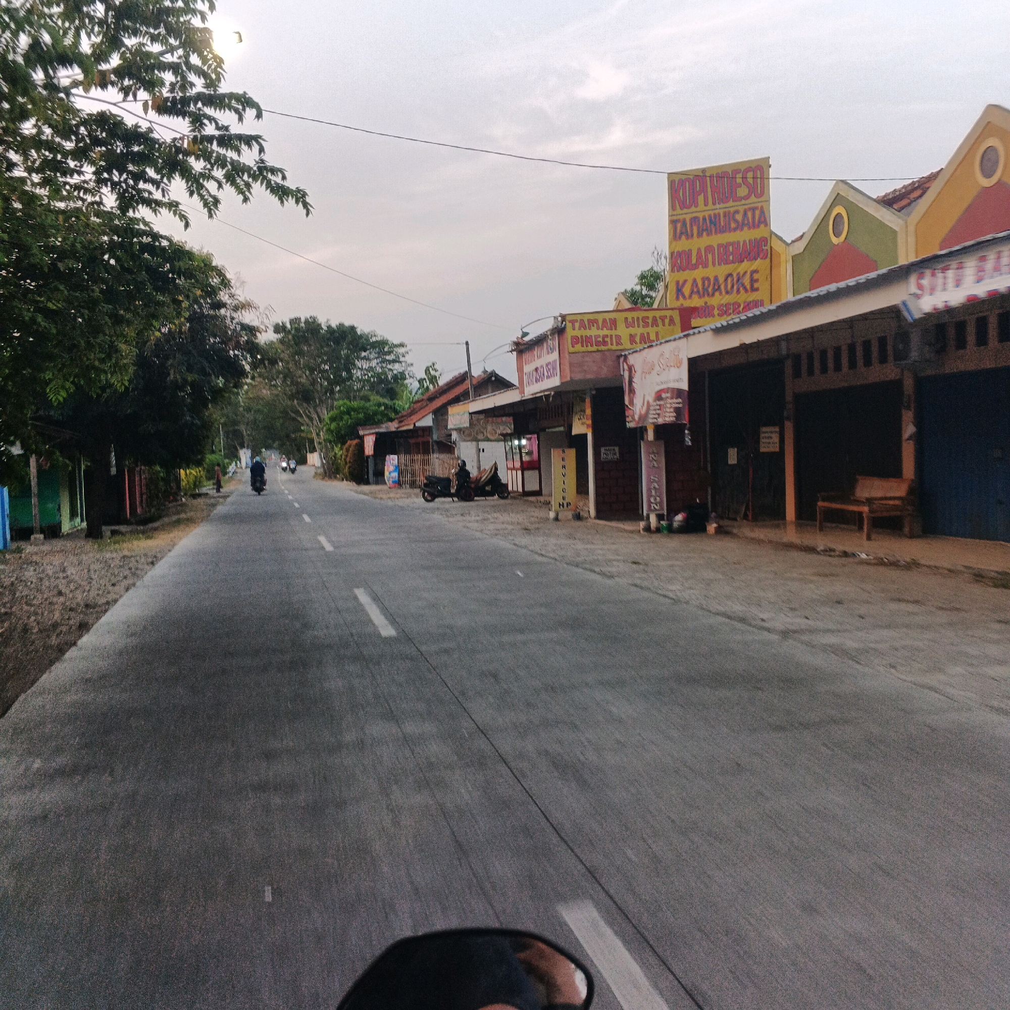 The image depicts a street scene in Desa Karangkemiri, Indonesia, captured from the perspective of someone riding a motorcycle. The road is paved with asphalt and marked with white lines. On the right side of the road, there are several storefronts with awnings. One storefront prominently displays a large yellow banner with red text that reads "KOPI HUESO TANAMAN WISATA KOLAM RENANG KARAOKE". Another sign above it reads "SOTO BA". Further down the street, there are more buildings and trees. On the left side of the road, there are trees and some small structures. In the distance, several motorcycles are visible on the road. The sky is overcast with a few scattered clouds, and the lighting suggests it is either early morning or late afternoon. A single bird is visible in the upper left portion of the sky.
