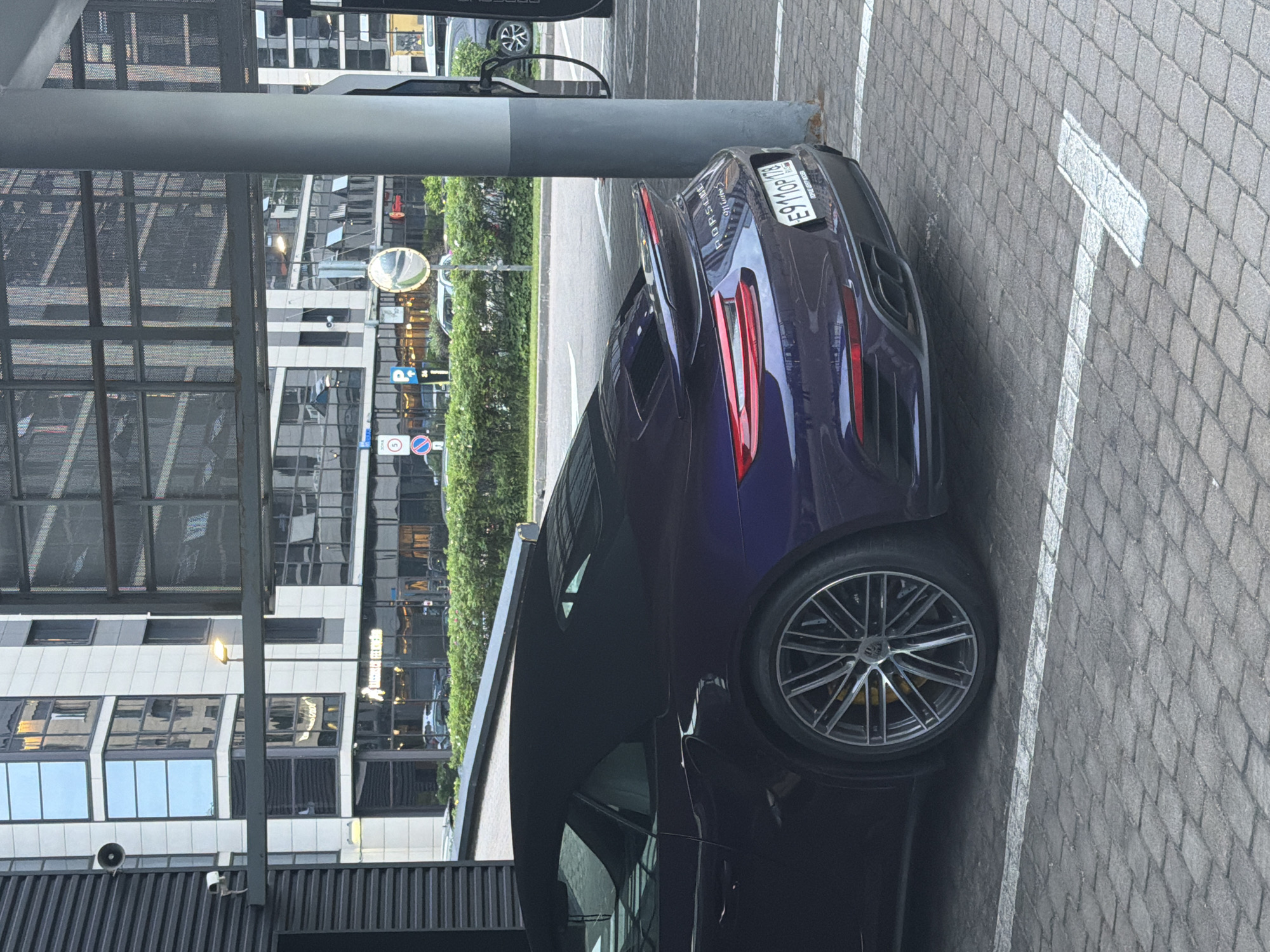 An eye-level, medium outdoor shot shows the rear passenger side of a dark purple Porsche 911 Turbo S convertible parked in a lot. The car is parked under a covered structure with a grey pillar to its right. The car's black convertible top is up. The license plate reads E911OP178. A charging cable is visible hanging from a unit attached to the pillar. The parking lot surface is paved with interlocking bricks and marked with white lines. In the background, a street with a row of green bushes separates the parking area from a modern building with many windows. Signs are visible on the street, including a circular sign with a red border and a cross inside, a blue parking sign, and a circular mirror. The lighting suggests it is daytime, possibly overcast.