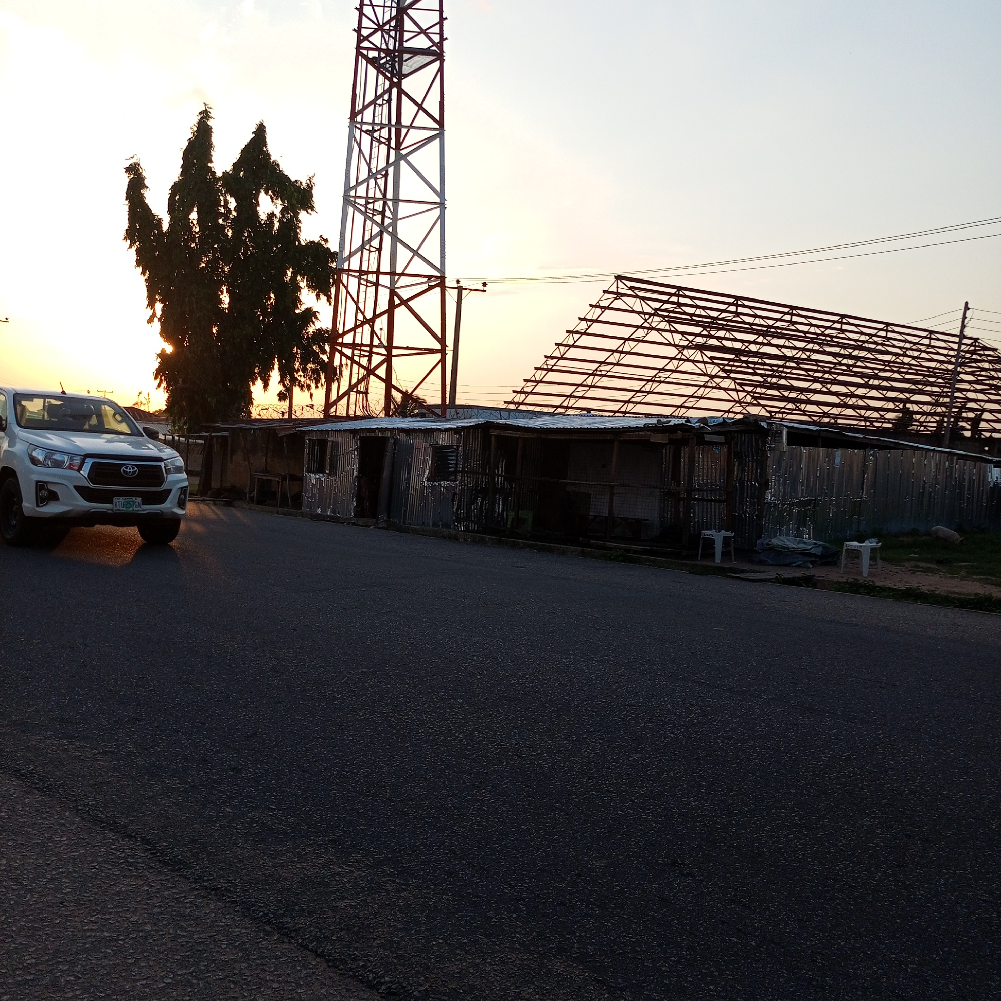 In Jalingo, Nigeria, a white Toyota Hilux pickup truck is parked on the left side of a paved road at dusk. The truck is facing right, with its headlights illuminated. Behind the truck, a large tree stands silhouetted against the sky, with a cell tower to its right. A low-lying structure made of corrugated metal, likely a roadside stall or workshop, is situated further down the road. The sky is a gradient of pale yellow and orange, indicating the setting sun. To the right of the stalls, the skeletal frame of a larger building under construction is visible, with its metal supports reaching towards the sky. The overall scene depicts a quiet street in a Nigerian city during the evening.