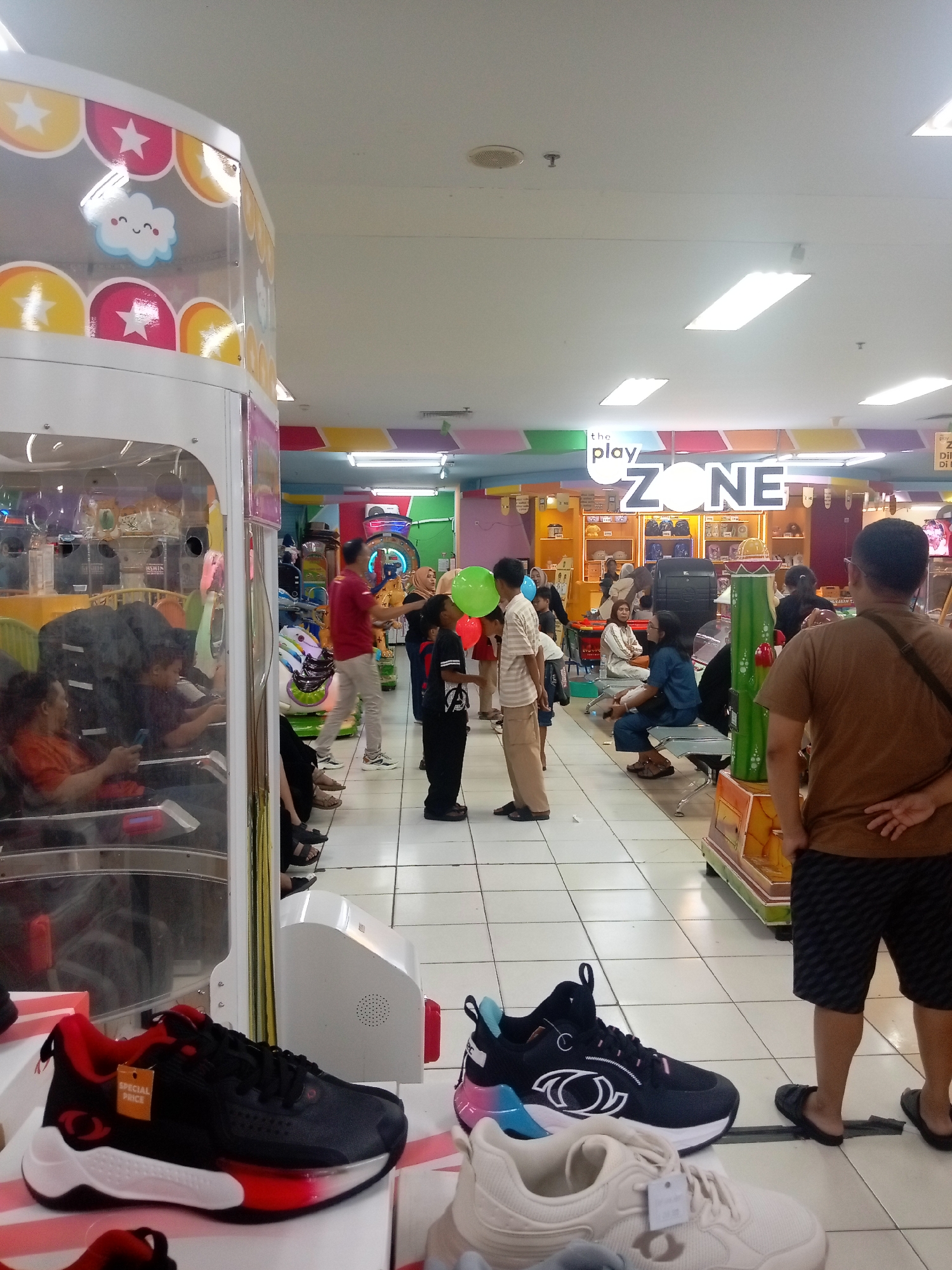 The image captures a scene inside what appears to be an indoor amusement area or arcade, likely located in Batam, Indonesia. In the foreground, there are several pairs of athletic shoes displayed on white boxes. One pair is predominantly black with red accents and a white sole, featuring a tag labeled "SPECIAL PRICE." Another pair, situated to the right, is a light beige color.
The midground reveals a collection of people, some standing and others seated. A man in a red shirt is visible, and further back, a group of people are gathered. One individual holds a green balloon, and another has a blue balloon. The signage in the background, notably "the play ZONE," suggests the area is a dedicated entertainment space for children or families. The environment features bright lighting from overhead fluorescent fixtures, a tiled floor, and colorful decorative elements on the walls and ceiling, including star and cloud motifs on a large arcade-style machine on the left. Several arcade games and rides are also visible.
The background shows more of the interior, with various attractions and seating areas. The overall impression is of a lively, family-friendly establishment.
