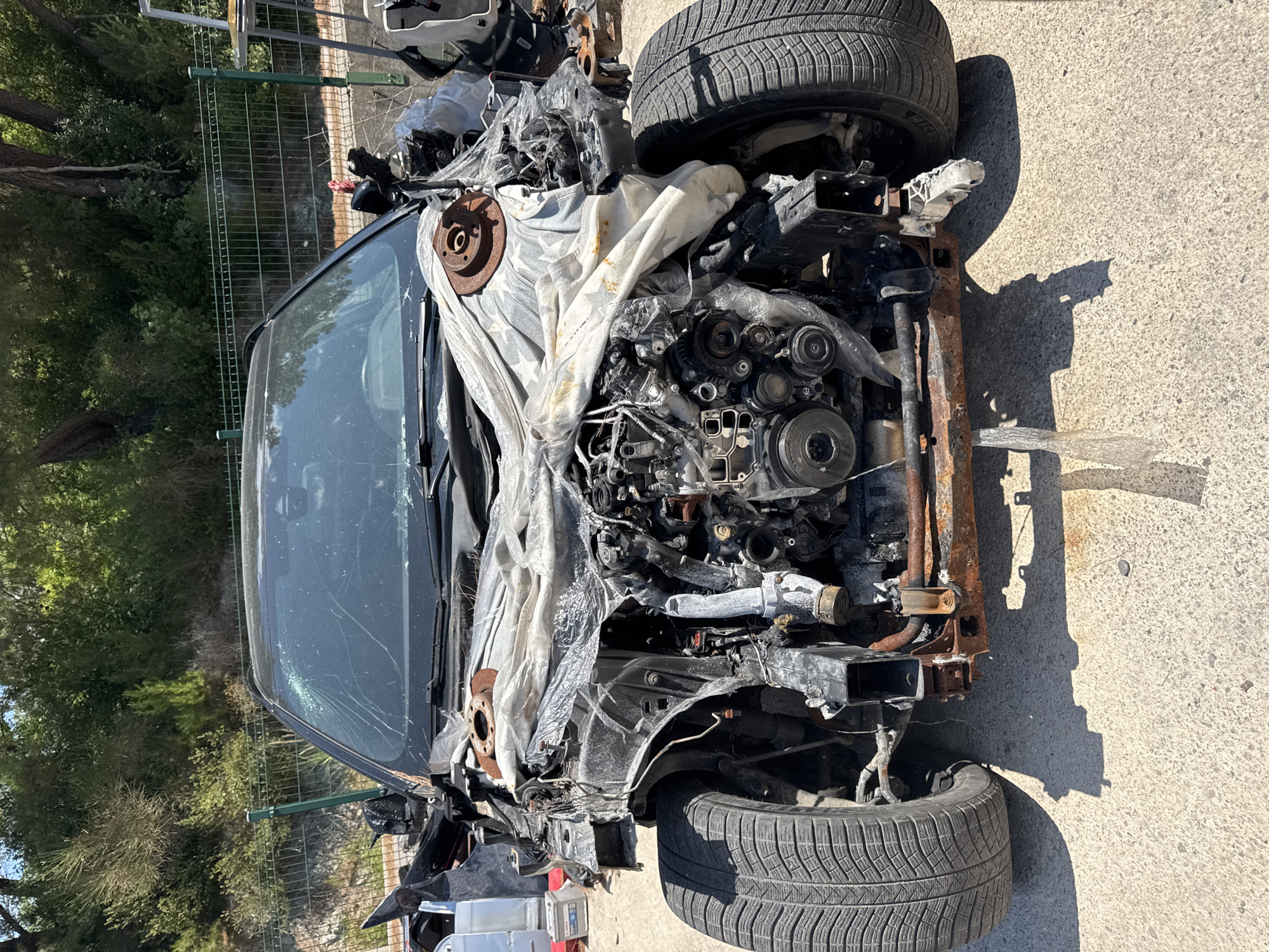 The image shows the front of a severely damaged car, likely abandoned, on a paved surface in Dub, Montenegro. The car's engine is exposed, with significant fire damage visible on its components and surrounding areas. The hood is partially covered by a white, scorched tarp or cloth, and the windshield is cracked. The front bumper is missing, and the car's body panels are warped and torn. Both front tires are still attached. The background includes a green metal fence, dense green foliage, and trees. The lighting suggests it is daytime, with strong shadows cast by the vehicle.