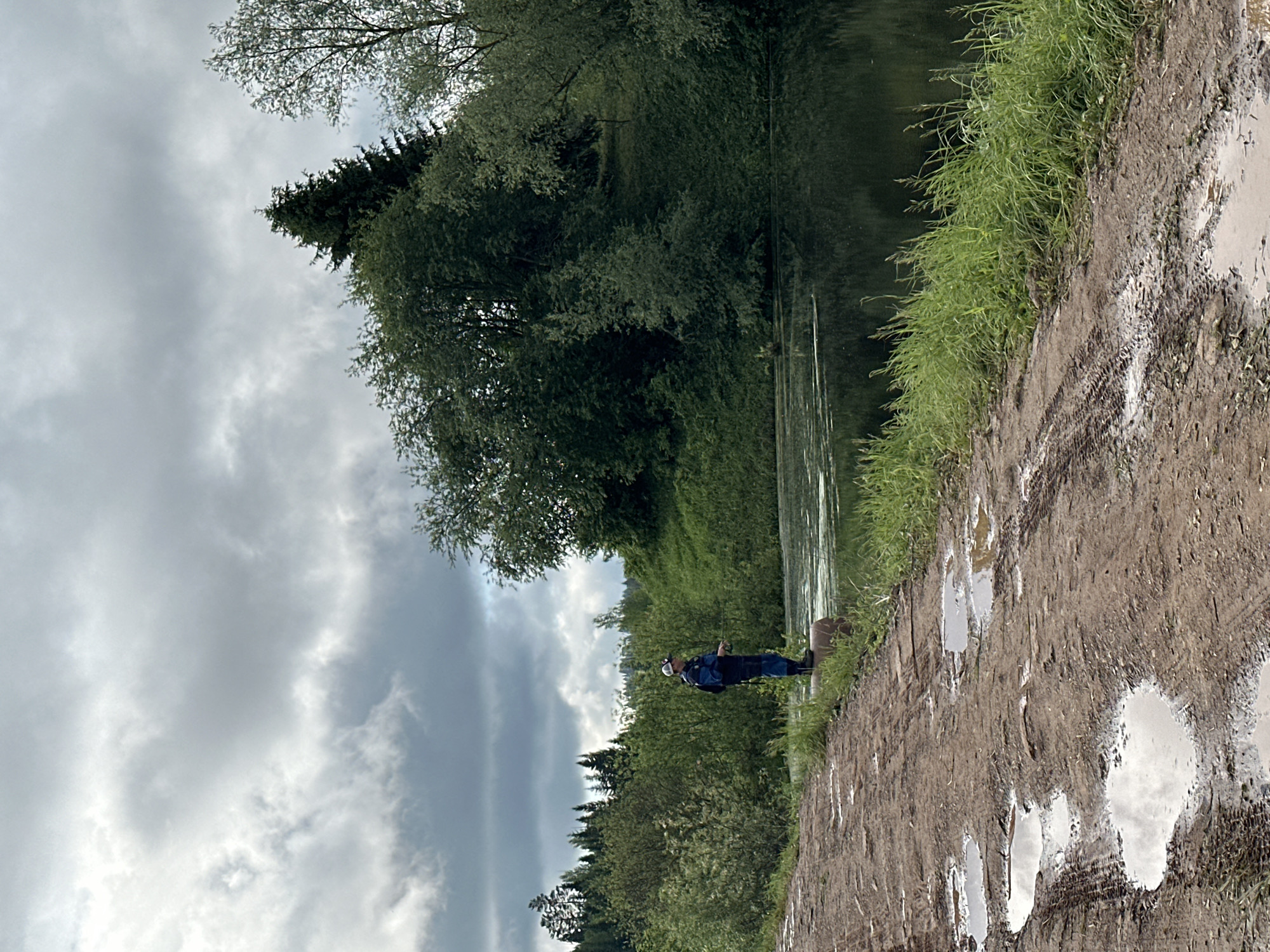 The image shows a person fishing along a muddy bank next to a body of water, likely a river or a lake. The location is indicated to be near Среднее Кечево, Russia. The ground is covered with tire tracks and standing water, suggesting recent vehicle traffic and rainfall. The person is standing near the edge of the water. The vegetation consists of grassy areas and trees along the water's edge. The sky is overcast.