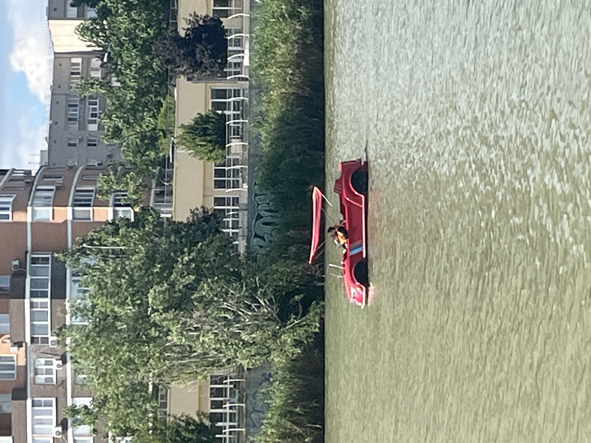 A bright red pedal boat, shaped like a race car, floats on a body of water. A person wearing an orange life vest is seated in the driver's position of the boat. The boat has a small sail or canopy. The water appears murky and greenish.
In the background, a lush green embankment rises from the water, densely covered with trees and bushes. Behind the greenery, several multi-story buildings are visible. These buildings have a consistent architectural style, featuring light-colored facades, numerous windows, and balconies. Some of the buildings appear to be residential apartment blocks.
The overall scene suggests a sunny day, likely during daylight hours, given the brightness of the scene and the shadows cast. The presence of the buildings and vegetation indicates an urban or semi-urban environment. The person in the pedal boat is engaged in a recreational activity on the water. There is no visible text in the image. The location context is Krasnodar, Russia.