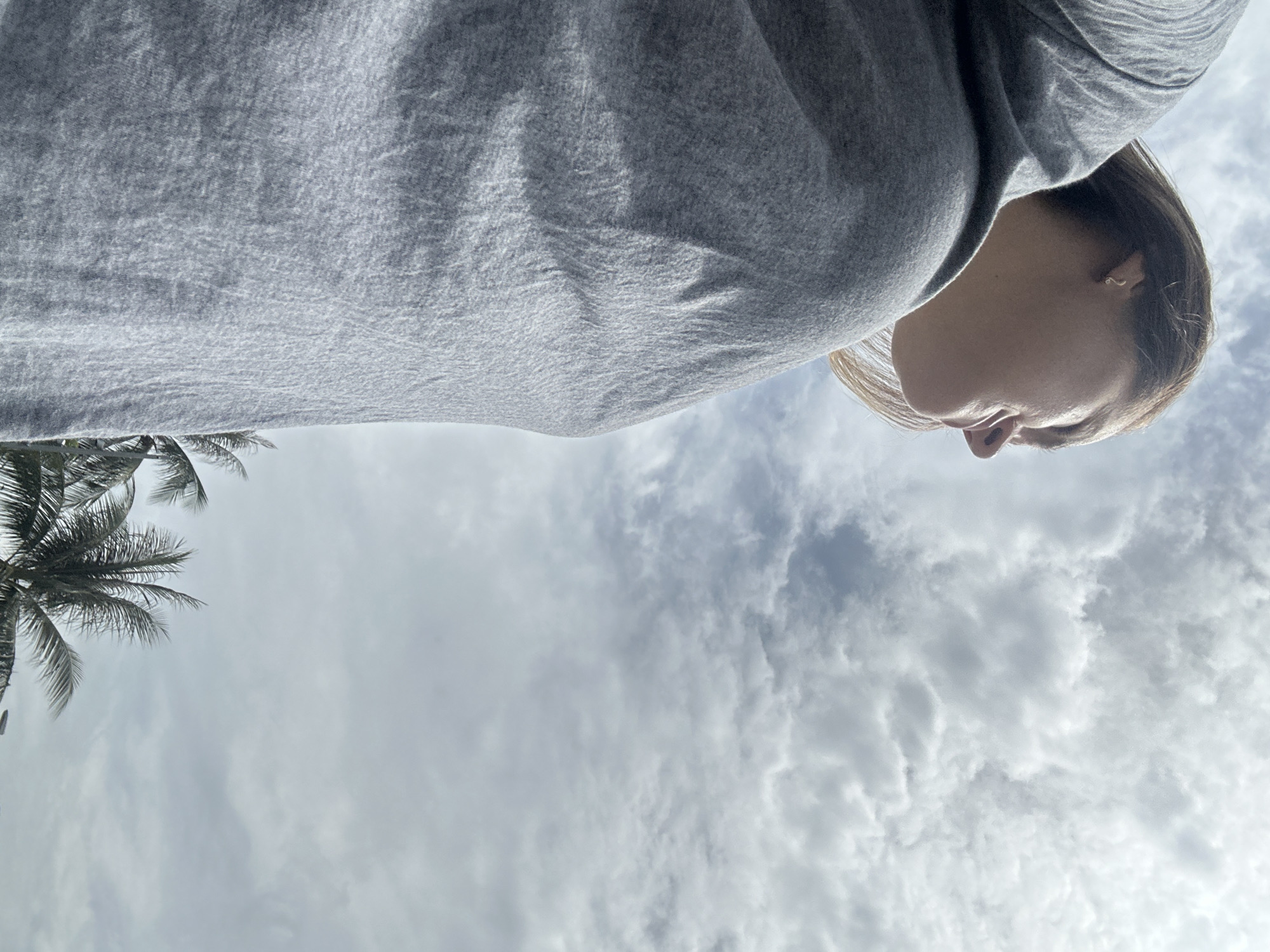 A low-angle shot captures a person wearing a grey top with their face visible. The background consists of a cloudy sky. A palm tree is visible in the lower left corner. The location is likely Bac Nha Trang Ward, Vietnam.