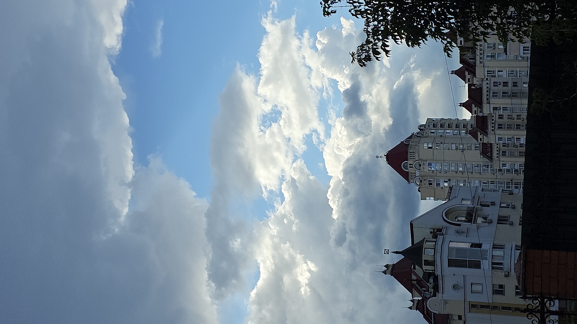 This image, captured in Kyiv, Ukraine, presents a view of several multi-story buildings against a dramatic sky filled with both bright blue patches and large, textured white and gray clouds. The architecture of the buildings is characterized by red-tiled, steeply pitched roofs, some adorned with spires. The central building is a tall, cream-colored structure with numerous windows and decorative arched elements near its peak, which is topped by a red roof and a small spire. To the left, a lighter-colored building features a distinctive turret with a conical roof and a circular window. Another building to the right echoes the architectural style of the main structures, with multiple red roofs. In the foreground, a dark fence, possibly made of wrought iron and brick, partially obscures the lower portion of the buildings. The upper right corner of the image includes the dark foliage of a tree, its leaves in shadow. The lighting suggests it is daytime, with sunlight illuminating parts of the clouds and the buildings, while other areas are in shadow. There are no people or vehicles visible in the frame.