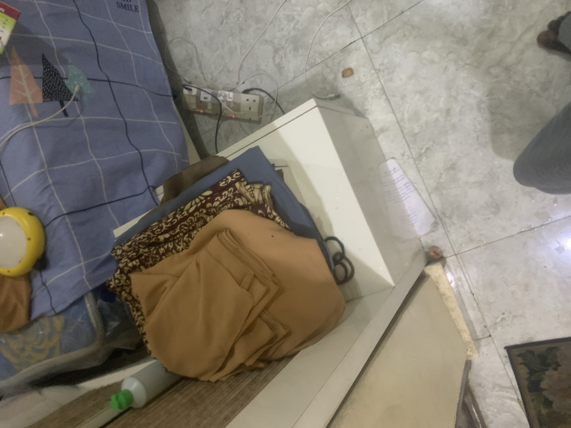 The image shows a room with tiled flooring. On the left side, there is a white cabinet with folded clothes on top, including a brown shirt and a patterned fabric. A green-capped bottle is also visible on the cabinet. A power strip with multiple outlets is connected to an electrical cord on the floor, with some plugs inserted. A blue blanket with a checkered pattern is spread on a surface behind the cabinet, with a yellow round object on it. In the background, a person's legs and feet are visible, indicating they are sitting or standing in the room. The overall impression is of a domestic setting, possibly a bedroom or living area, in Kuson, Nigeria.