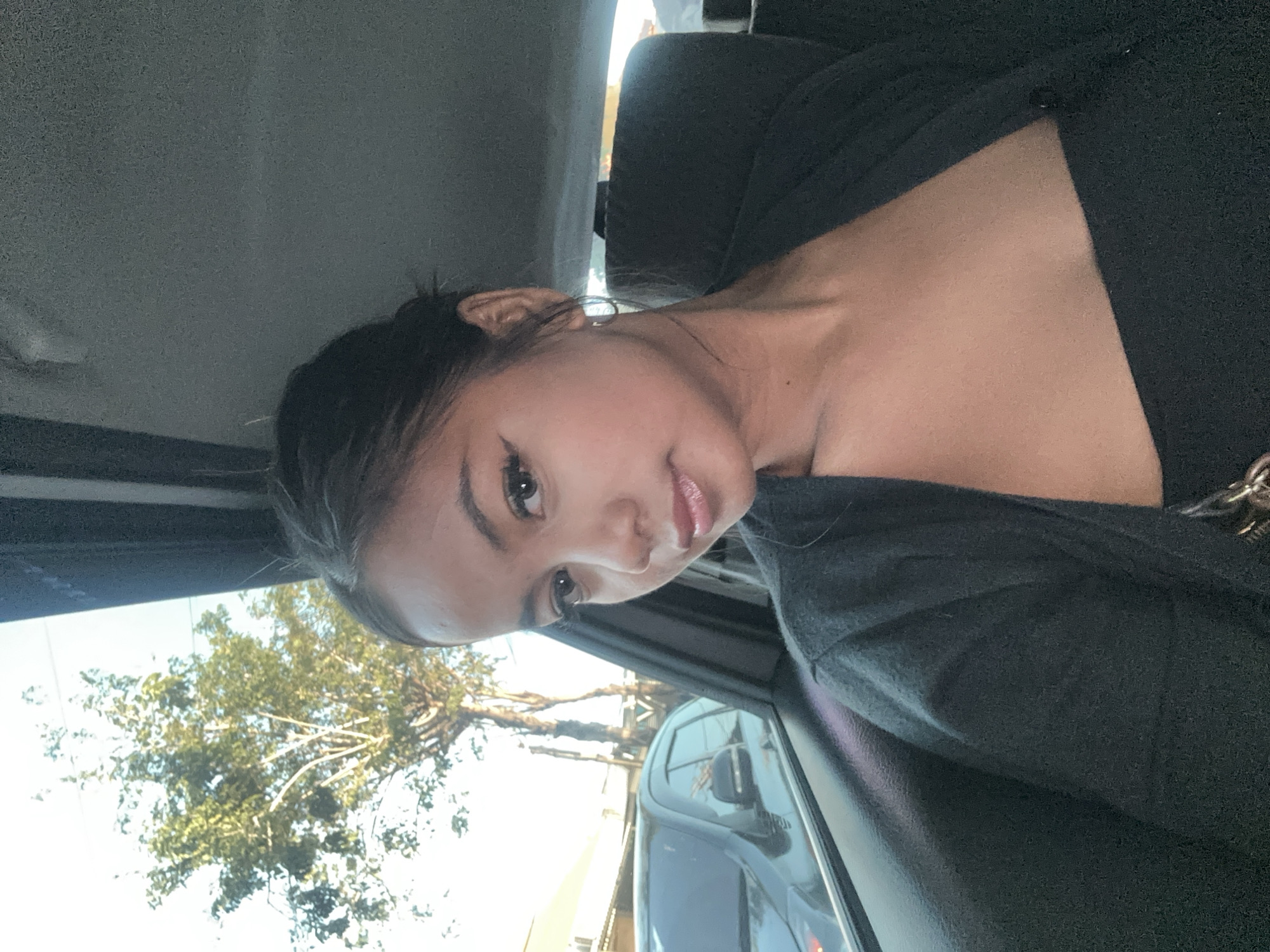 A young woman with dark hair pulled back is sitting in the backseat of a car. She is looking towards the camera with a slight smile. She is wearing a black top and a dark cardigan. The background shows the interior of the car and out the window, where a tree and a street with buildings are visible. The scene appears to be in North Kuta, Indonesia, given the context provided.