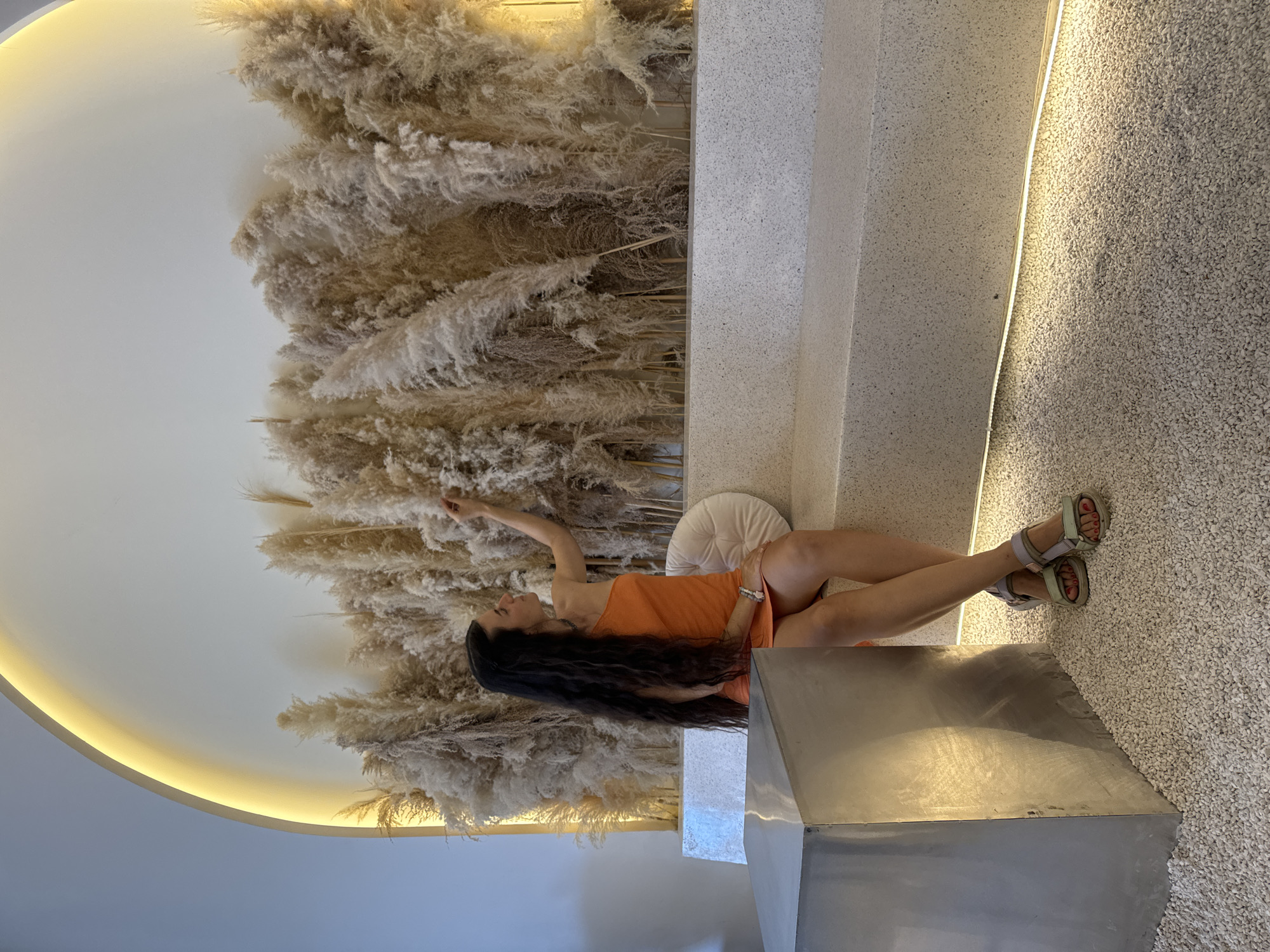 A woman poses in a modern, artfully designed indoor space located in Phu Thuy Ward, Vietnam. She is seen lying on her side on a light-colored, speckled concrete bench or ledge, propped up on her left elbow. Her long, dark hair flows down past her shoulders, and she wears an orange sleeveless top or dress, paired with light-colored sandals that reveal red nail polish on her toes. A subtle bracelet is visible on her left wrist. Her right arm is extended upwards, while her left hand rests on her thigh, and her gaze is directed upwards, conveying a relaxed and thoughtful demeanor.
The backdrop is a striking wall entirely covered with feathery, dried pampas grass in varying shades of beige and light brown, creating a soft, textured visual. Behind her, a round, light-colored cushion adds to the comfort. To the left of the frame, a curved white wall is illuminated by warm yellow backlighting, enhancing the contemporary aesthetic. The floor is covered with small, light-colored pebbles, and a reflective metallic, geometric object sits in the foreground. The indoor lighting suggests a controlled environment, possibly during the evening or within a cafe or showroom. No visible text is present.