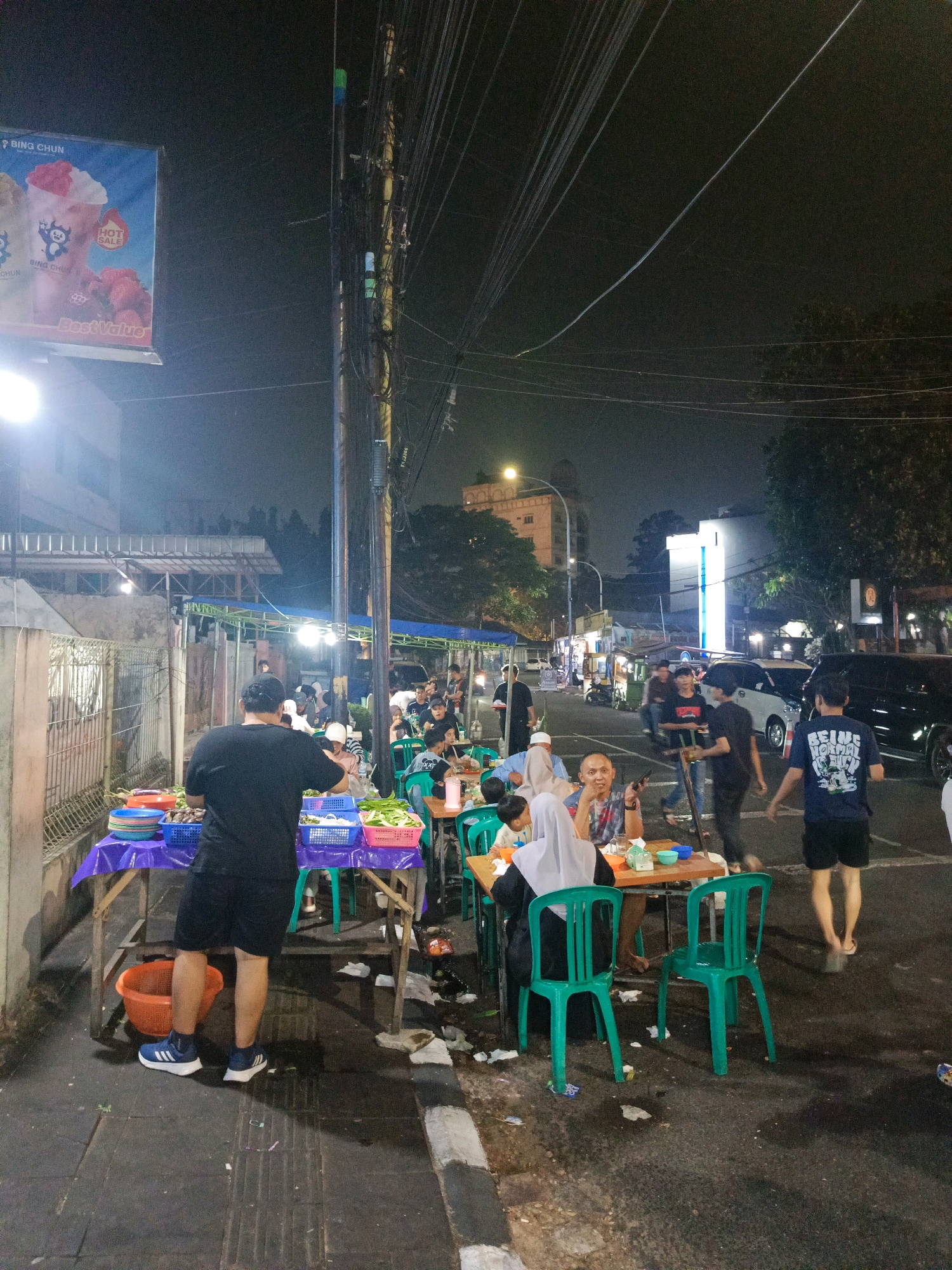 The image depicts an outdoor night scene at a bustling street food market, likely in Tangerang, Indonesia, characterized by food stalls and diners. In the foreground, a man in a black t-shirt and shorts stands behind a table laden with plastic containers of food, including what appears to be vegetables and fruits. He is wearing blue and white Adidas sneakers. To his right, several green plastic chairs are arranged around tables where people are seated, eating and socializing. Some diners are wearing head coverings, suggesting a diverse clientele. The background reveals a street with passing vehicles, including cars and motorcycles, and buildings lining the road. Overhead, a complex network of electrical wires crisscrosses the frame, illuminated by streetlights and the glow from the stalls. The ground is paved and shows signs of litter, typical of a busy public area. A large billboard advertising "Bing Chun" with images of ice cream and strawberries is visible on the left side of the image. The overall atmosphere is one of a vibrant, casual dining experience in a lively urban environment.