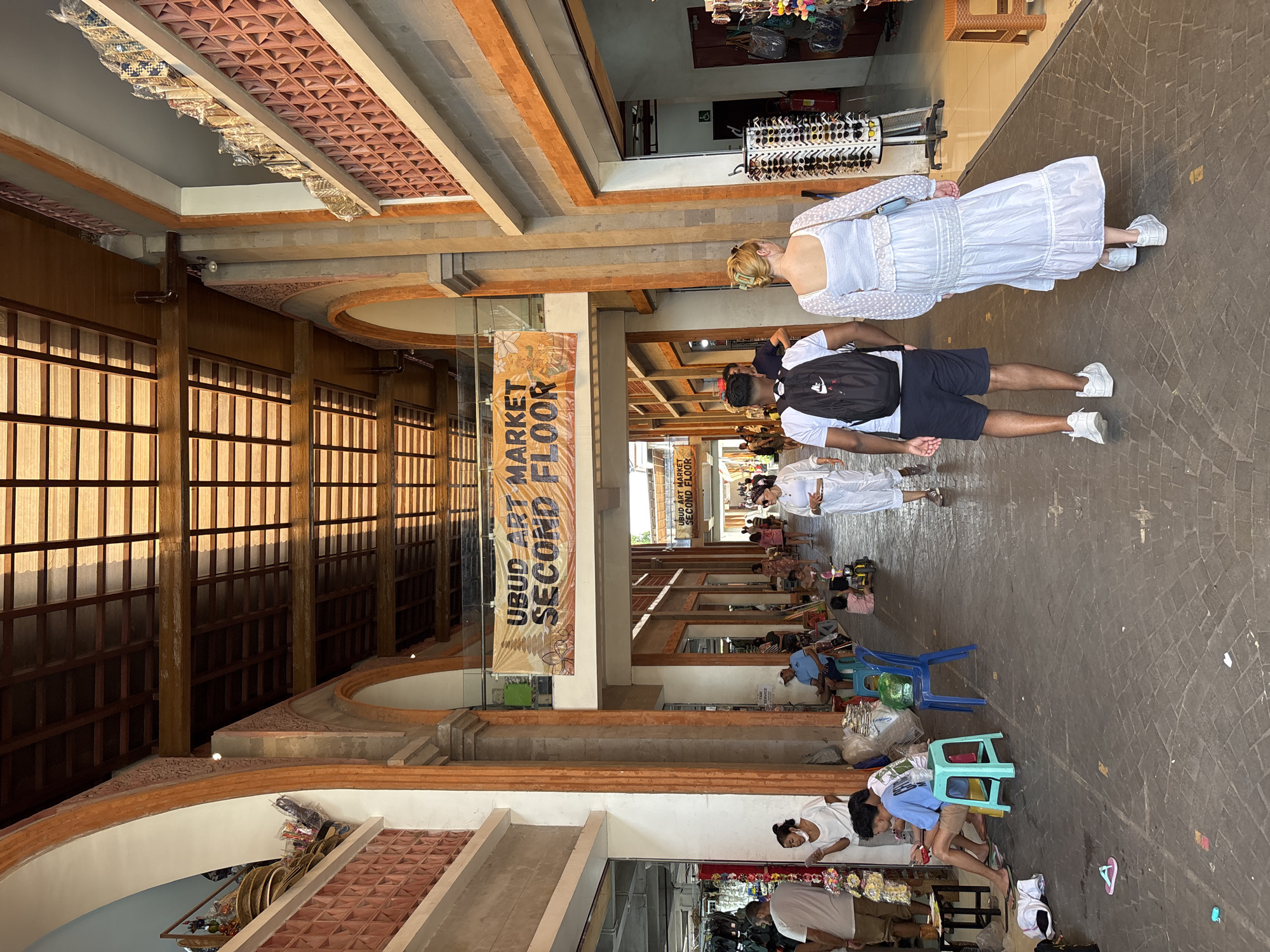 The image shows the Ubud Art Market Second Floor in the Ubud District, Indonesia. The scene depicts an indoor marketplace with vendors sitting on low stools near their wares. Shoppers, including a couple, are seen walking along the corridor. The architecture features arched structures and a terracotta-tiled ceiling. A large banner reading "UBUD ART MARKET SECOND FLOOR" is displayed prominently. The marketplace is bustling with activity.