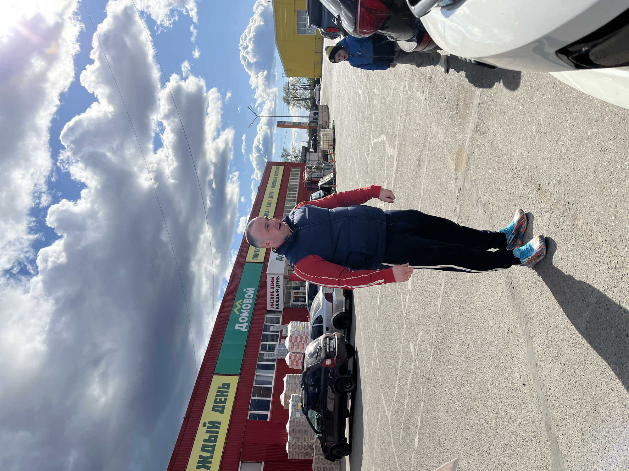 A full-length image shows a man standing outside the “Домовой” (“House”) store in Chaikovsky, Russia. The man wears a navy vest over a red jacket and black pants with a white stripe. He wears bright, patterned sneakers. The store building is red with green and yellow signage. A parked black car and stacks of bagged materials are near the store entrance. Another person stands by cars near a yellow building. The sky is partly cloudy.