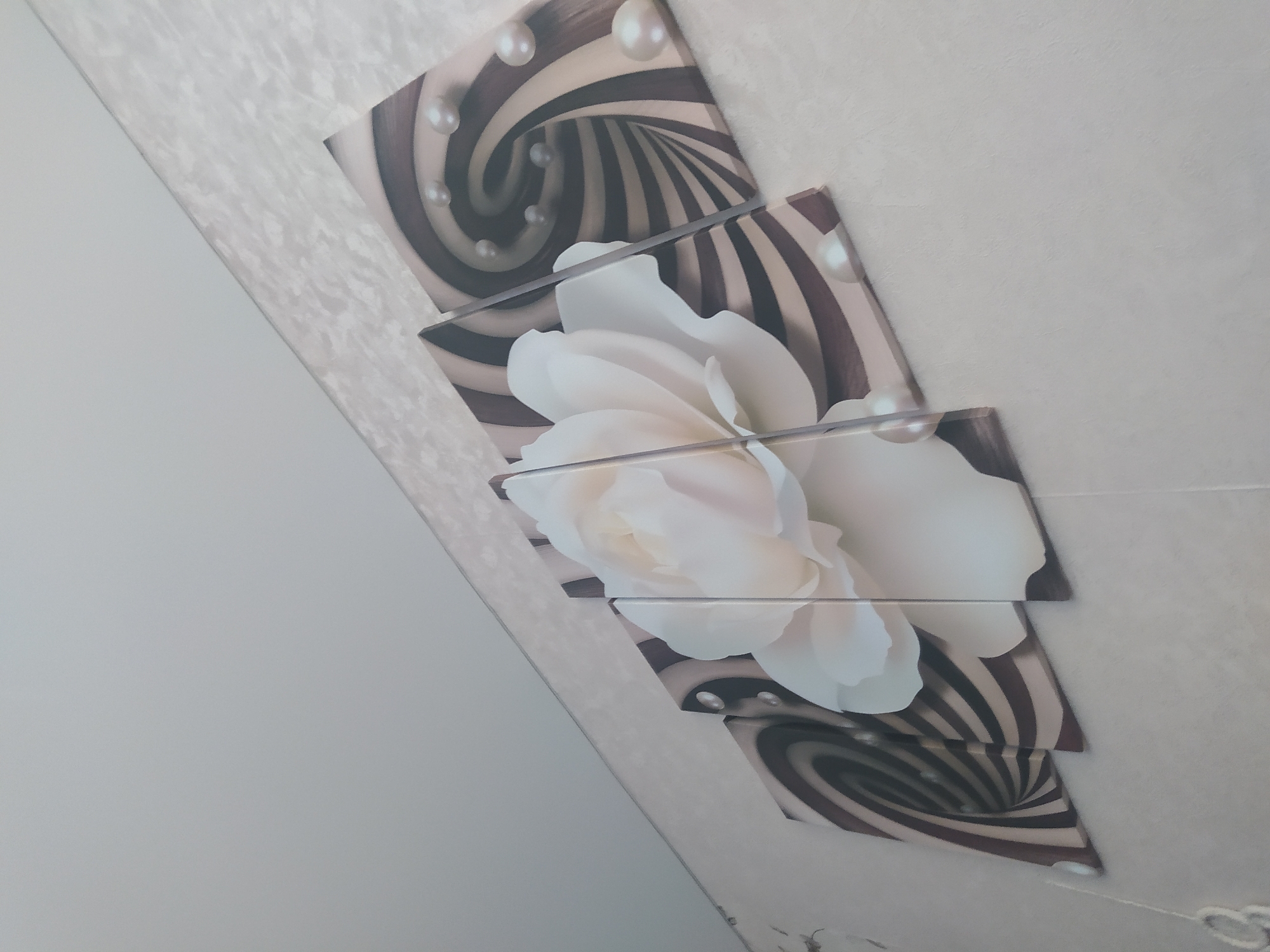 The image shows a four-panel canvas art piece hanging on a textured, light-colored wall. The artwork features a large, white rose in the center, with swirling, abstract patterns in shades of brown and beige on the outer panels. Several pearls are depicted scattered across the artwork, some appearing to be embedded within the swirling patterns. The room appears to be an indoor setting, possibly a home, with a plain white ceiling visible at the top left of the image. The specific location of Gusev, Russia is not discernible from the visual information provided in the image.