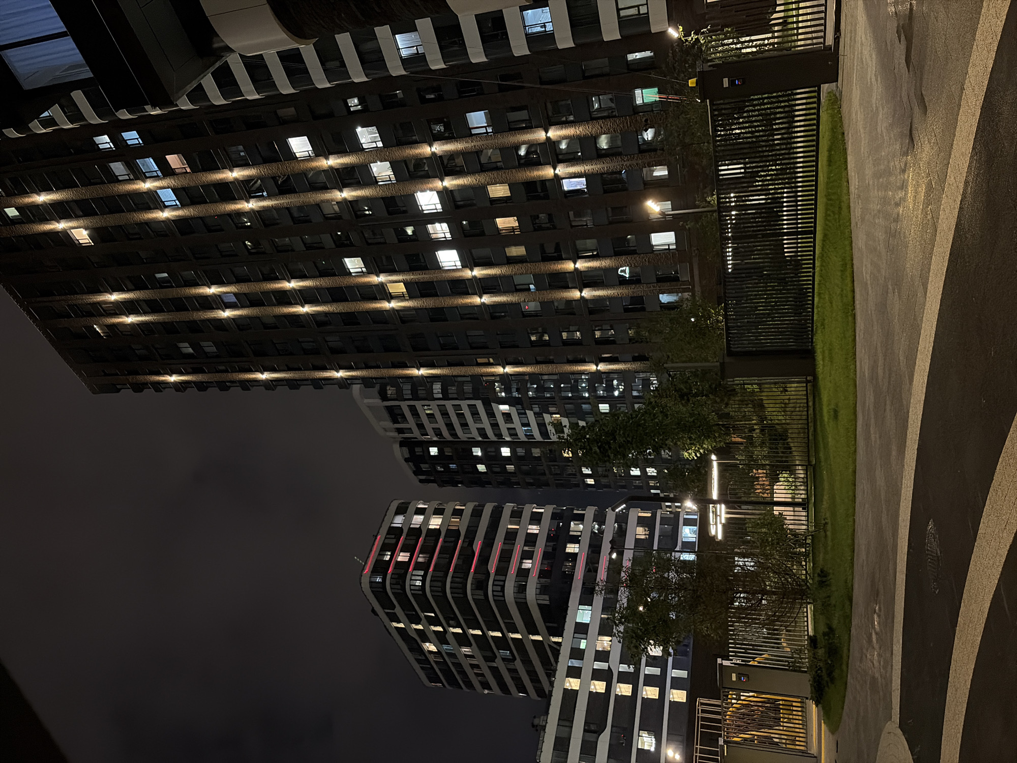 This image captures a nighttime view of a modern residential complex in Moscow, Russia. The scene is dominated by several tall, multi-story buildings with numerous illuminated windows, suggesting occupancy. Vertical lighting fixtures on the facades of the buildings highlight their architectural details. In the foreground, a paved walkway with white markings curves around a grassy area enclosed by a metal fence. A gate is visible on the right side of the frame. Trees and shrubbery are present within the fenced area, adding a touch of greenery. The sky is dark and cloudy, indicating nighttime. The overall impression is one of a well-lit, contemporary urban living environment at night.