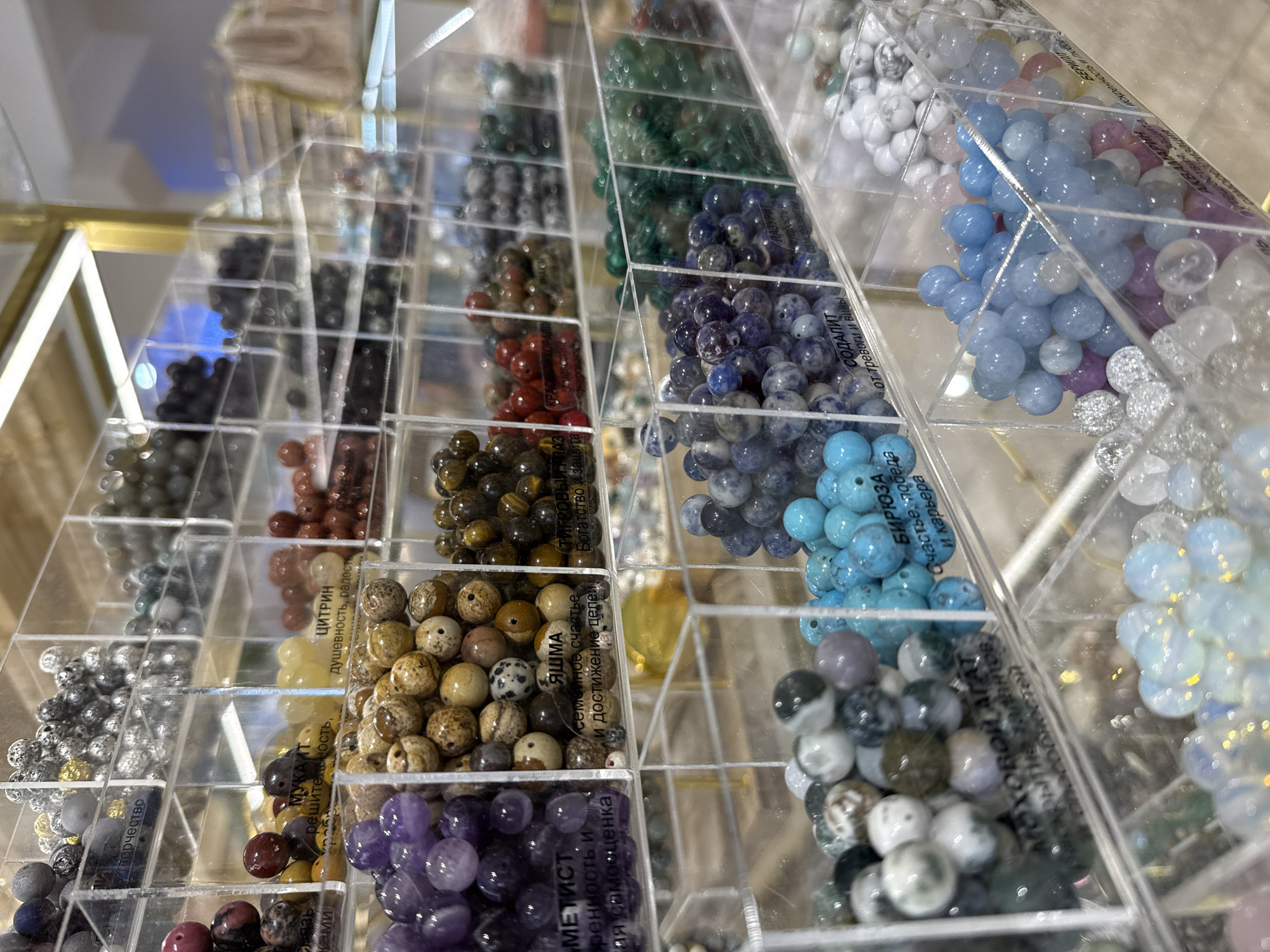 The image displays a retail display of various gemstone beads organized in clear plastic compartments arranged on shelves. The beads vary in color, size, and texture, including shades of purple, brown, yellow, red, green, blue, gray, and white. Some compartments have labels with Russian text, suggesting the beads are identified by type and possibly their purported properties. The display is illuminated by overhead lighting, creating reflections on the plastic surfaces. In the background, a light-colored bag and part of a golden display structure are visible. The overall environment appears to be an indoor retail space, likely a jewelry or craft supply store. The style of the display and the presence of Cyrillic labeling suggest this scene is located in Saint Petersburg, Russia. No individuals are clearly depicted in the image.