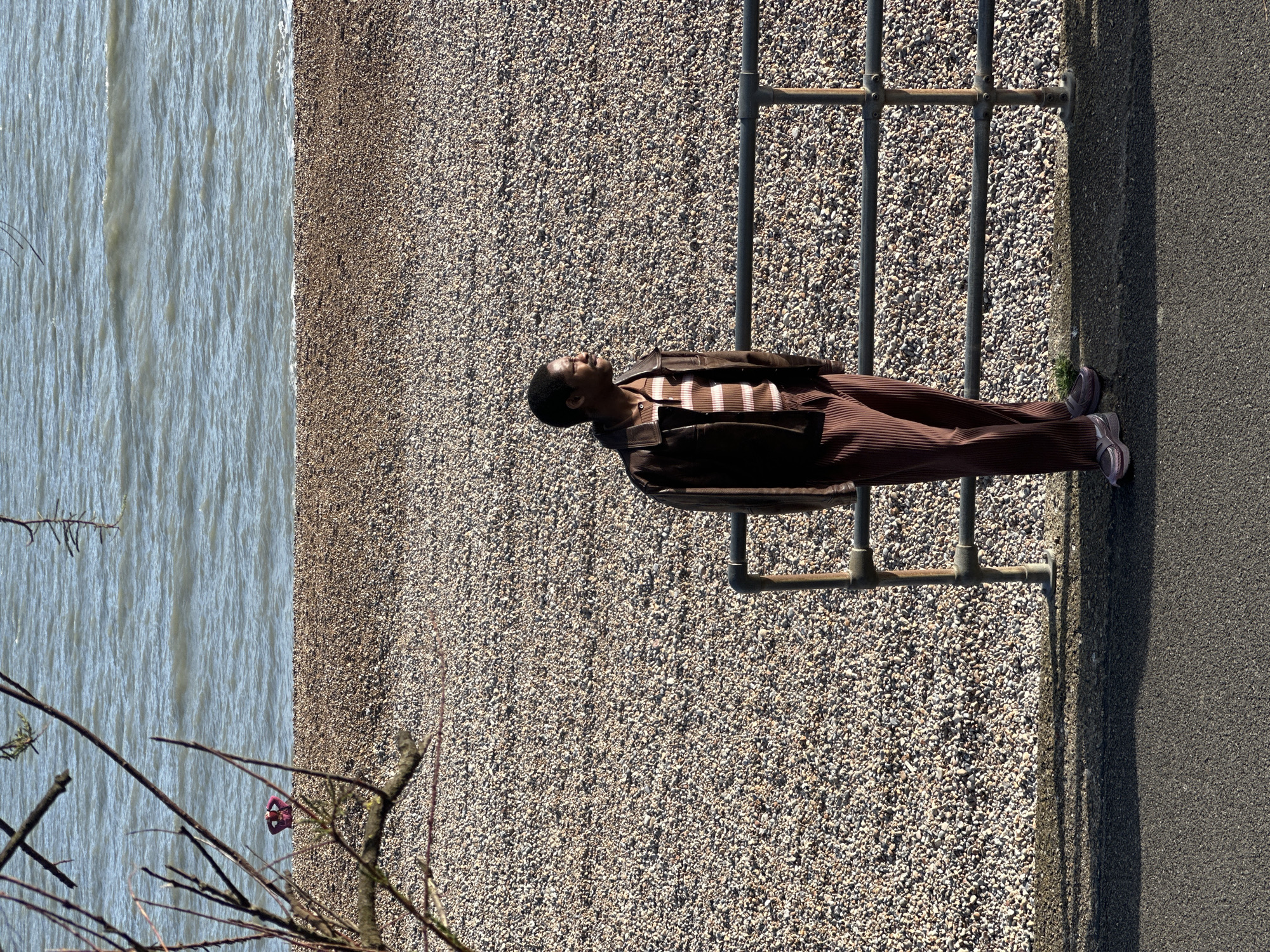 A man lies horizontally across a metal railing dividing a pebble beach from a paved walkway. The beach extends to the left of the frame, bordered by a body of water. The man wears a dark brown jacket, a striped brown and white shirt, and brown pleated trousers. His shoes are light-colored. Branches of a tree are visible in the lower left corner. The scene is set in Folkestone and Hythe District, United Kingdom.