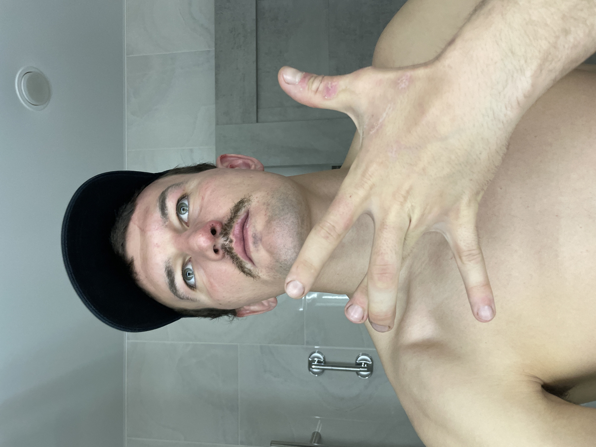 A person with a dark cap and a drawn-on mustache is posing indoors in front of a tiled wall. The person has light skin and light-colored eyes. Their hand is raised, obscuring part of their chest. The scene appears to be a bathroom, judging by the tiled walls, a grab bar, and a ceiling fixture. The location is unspecified and the claim of being in Sochi, Russia, cannot be substantiated based solely on the visual information provided.