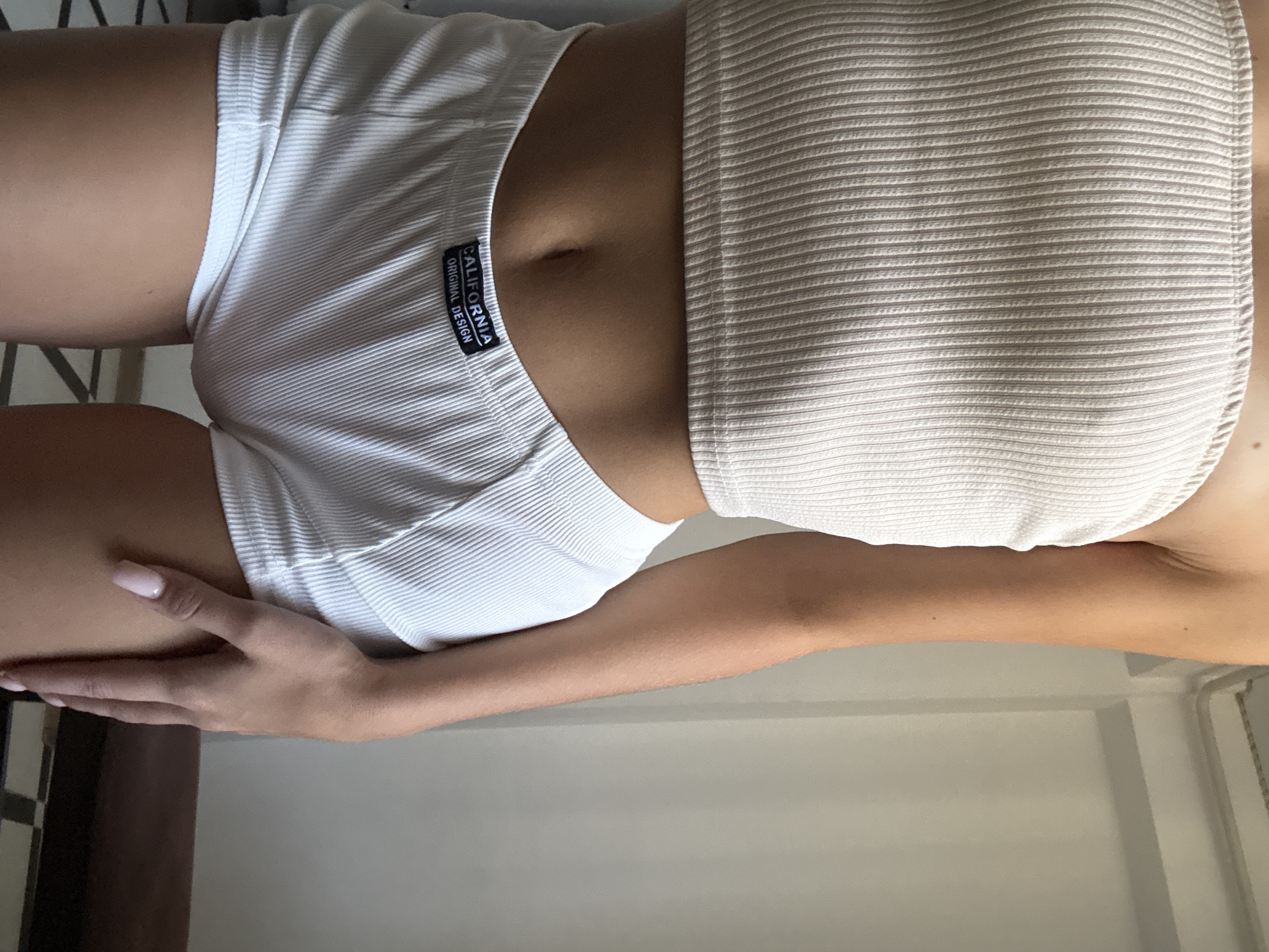 A cropped, medium shot shows the midsection of a person wearing a beige ribbed bandeau top and white ribbed boy shorts. The shorts have a black label on the front that reads "CALIFORNIA ORIGINAL DESIGN." The person's skin is tan, and they have light pink nail polish on their fingernails. The background is a plain light-colored wall with some molding at the top, and a dark brown surface is visible at the bottom right. The floor appears to be tiled with a black and white checkered pattern. The image does not provide enough information to determine the location in Chișinău, Moldova.