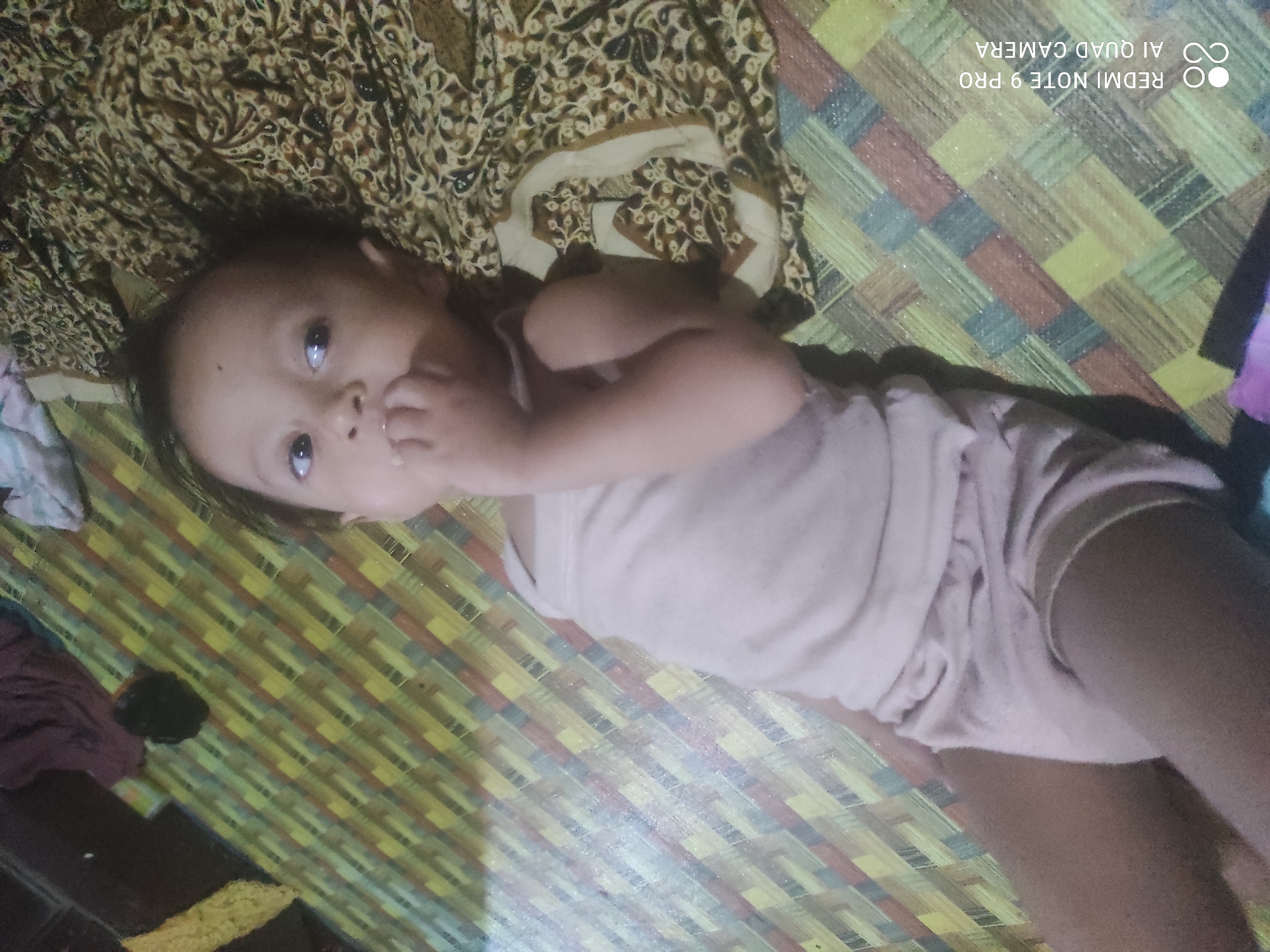 The media file is a photograph taken in Sakra, Indonesia, depicting a young child lying on their back on a woven mat. The child is wearing a light-colored tank top and shorts. Their head is tilted slightly, and their eyes are open, looking upwards. One hand is near their mouth, suggesting they might be sucking their thumb or exploring. The background features a patterned fabric and the woven mat has a diagonal design of yellow, red, and grey stripes. The lighting suggests it is daytime. Visible text in the upper right corner reads "REDMI NOTE 9 PRO AI QUAD CAMERA".