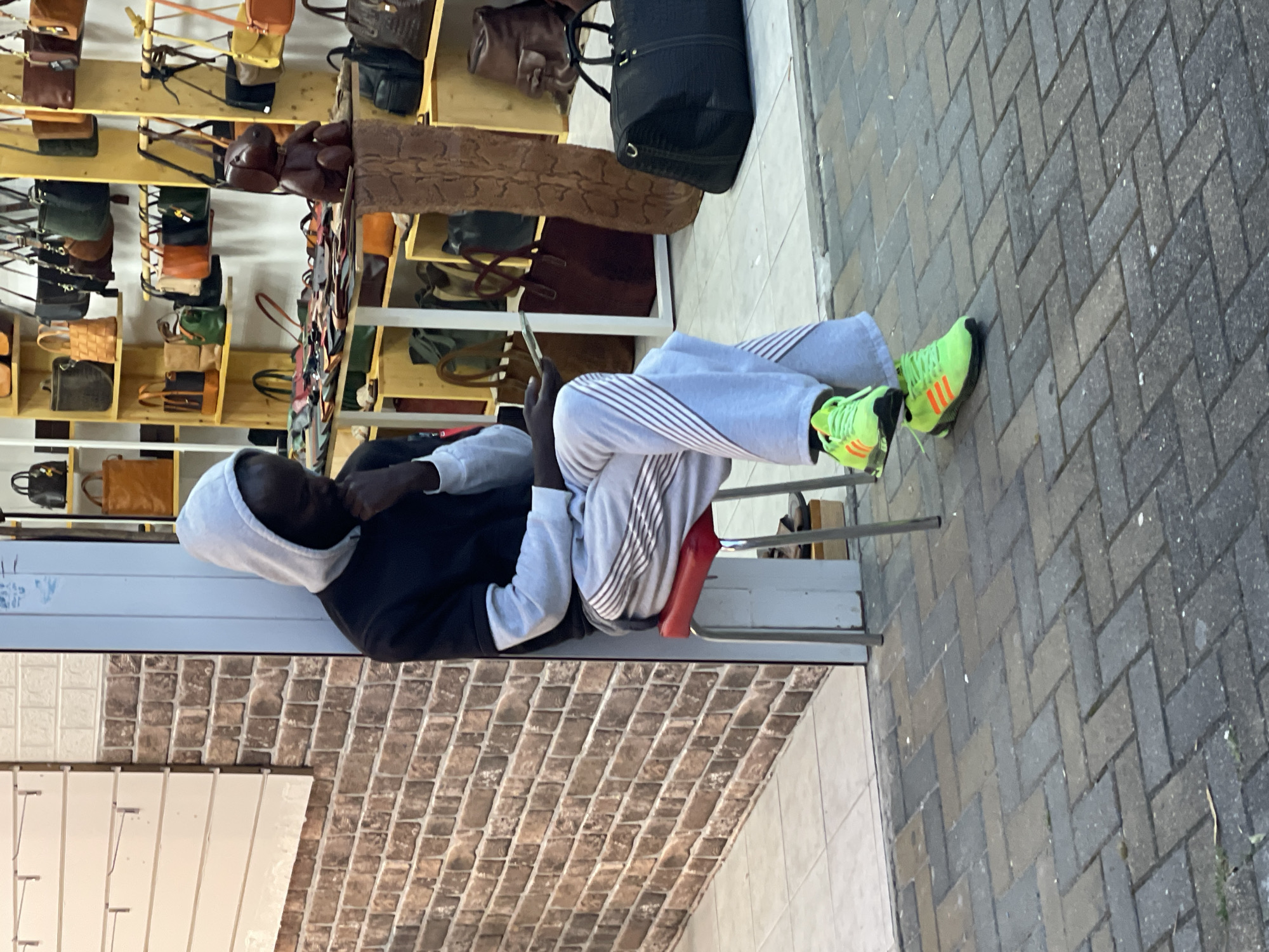A person is sitting on a red chair on a sidewalk in Sochi, Russia, looking at a phone. The person is wearing a black hoodie, grey striped sweatpants, and bright green sneakers with orange stripes. Behind the person is a brick wall, a white wall, and shelves displaying purses and bags. The sidewalk is paved with grey bricks.
