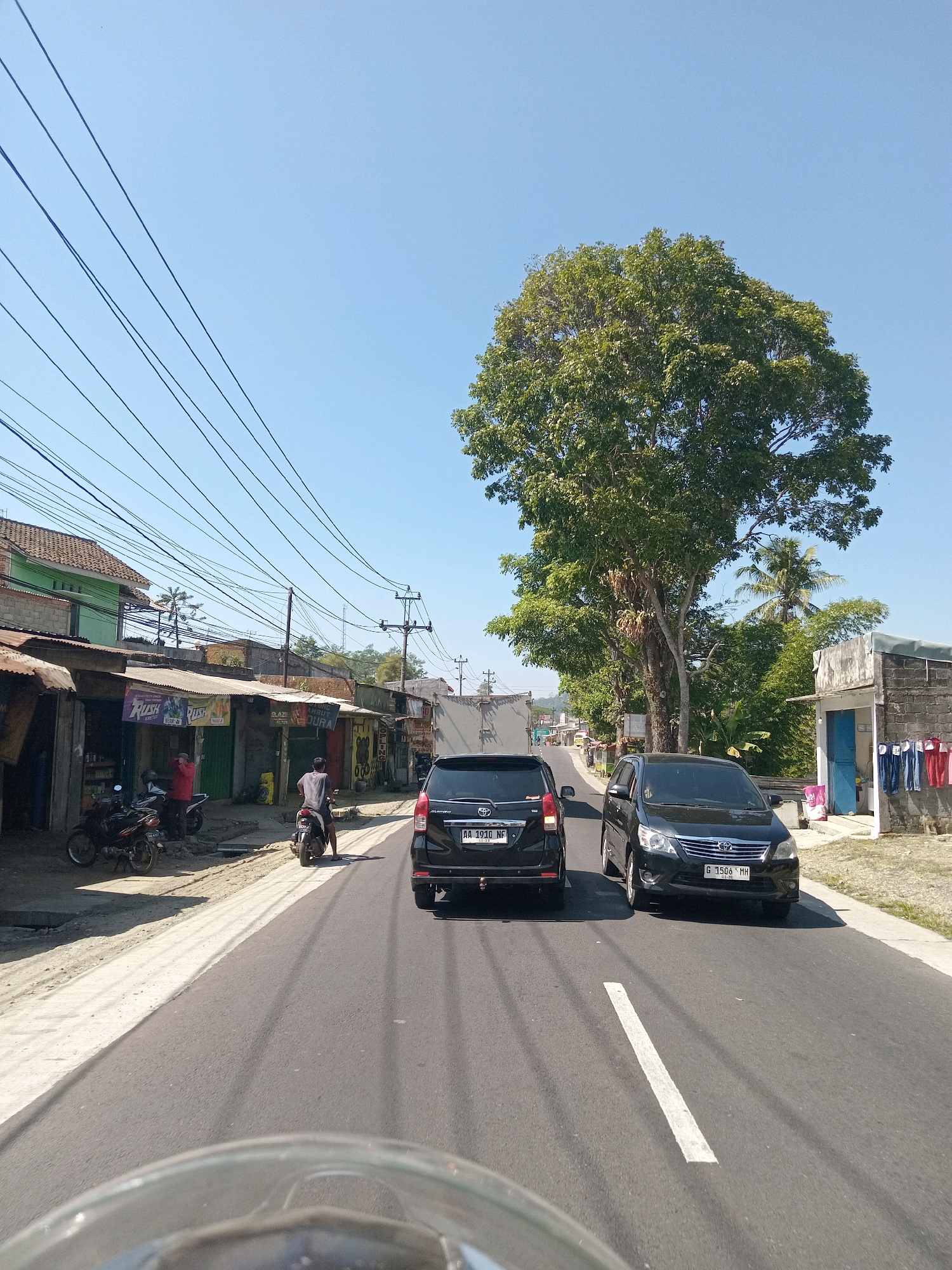 This is a horizontal, eye-level, medium shot of a rural road in Maduretno, Indonesia, on a bright, clear day. The foreground is dominated by the reflective edge of what appears to be a motorcycle's windshield. Two black cars, a Toyota Avanza and another similar model, are traveling on the asphalt road, which has a white dashed line dividing the lanes and a solid white line marking the shoulder. A single motorcyclist, wearing a grey shirt and dark shorts, is seen from the rear, also on the road. To the left of the road, there is a row of small shops with corrugated metal roofs, displaying various signs, including "RUSH." Several motorcycles are parked in front of the shops, and two people are visible near them, one wearing a red shirt. The background features a large, leafy green tree to the right of the road, and on the far right, a simple building with a blue door and clothes hanging on a line. Utility poles and numerous power lines stretch across the upper portion of the image, against a clear blue sky. The overall scene depicts a typical street in a developing area, with a mix of commercial activity and everyday traffic.