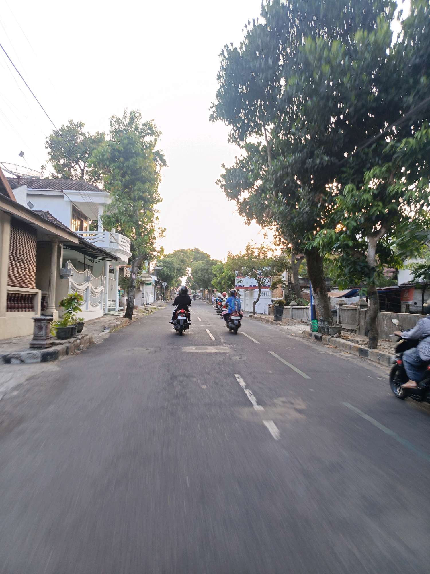 The image captures a street scene in Magetan, Indonesia, likely during daylight hours, possibly late afternoon given the soft lighting. The perspective suggests the photo was taken from a moving vehicle, possibly a motorcycle, looking forward down a paved road.
On the left side of the road, there are houses with white walls and tiled roofs. One house has a distinct white balcony and decorative railings. Potted plants and a stone planter are visible along the sidewalk. Trees with dense green foliage line the road, creating shade.
Several people are riding motorcycles. In the foreground on the right, a person is riding a motorcycle, wearing a light-colored top and blue jeans, with their feet visible. Further up the road, two motorcycles are visible side-by-side, with riders wearing helmets. One rider has a blue backpack. Another motorcycle is further ahead, with a rider in black.
The road has white markings, including a dashed white line. The environment appears to be a residential or mixed-use area within the city, characterized by the presence of homes, trees, and ongoing vehicular traffic. The sky is a bright, overcast white.