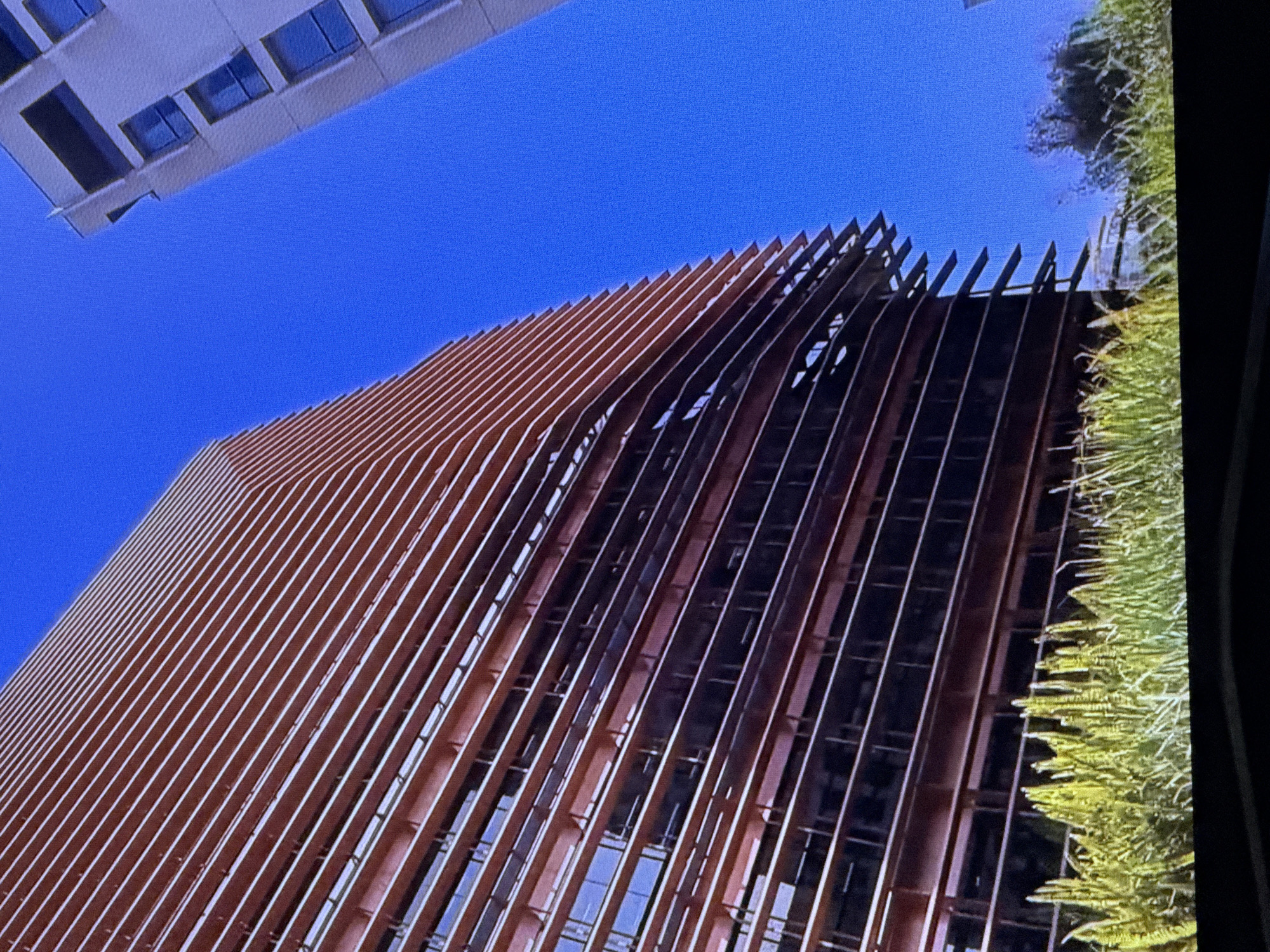 An exterior, low-angle, close-up shot captures the facade of a modern copper-colored building with horizontal louvers, contrasted against a bright blue sky. The building's design features repeating horizontal bands that extend outwards, creating a layered effect. To the right of the frame, another contemporary building with a white facade and dark-tinted windows is partially visible. In the foreground, lush green grass and foliage are prominent. The scene is devoid of any people or visible actions. The architectural style and the clear blue sky suggest an urban setting in Dubai, United Arab Emirates.