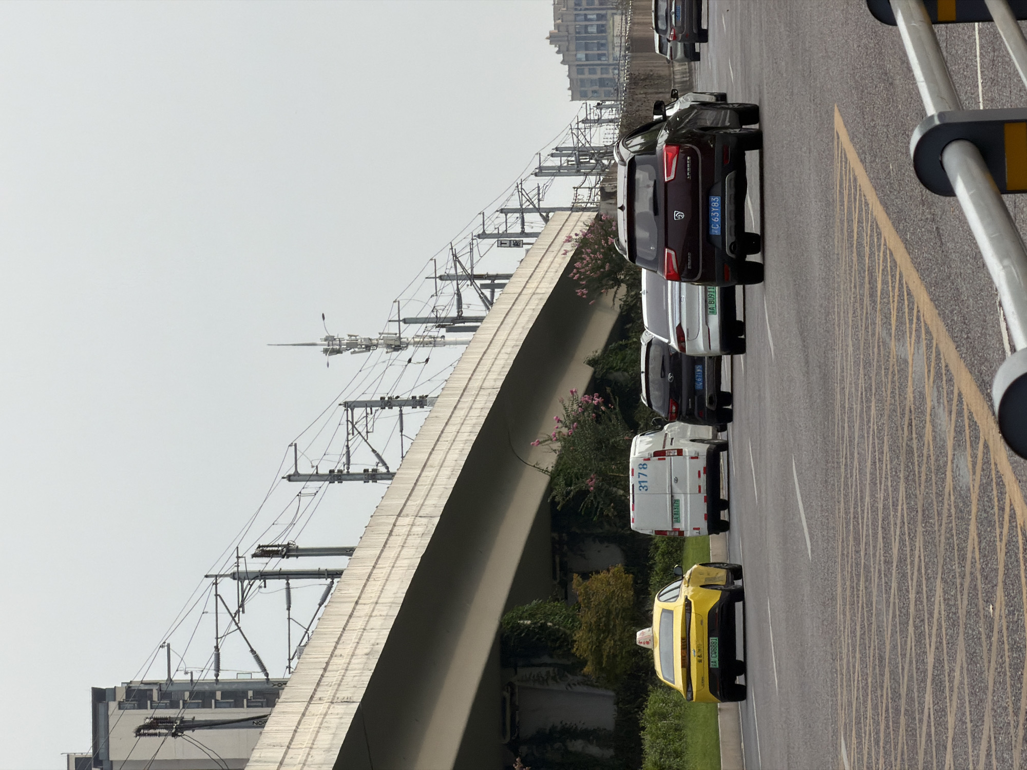 In Jiangjin, China, a yellow taxi with license plate "A CQS881" is positioned in the left lane of a multi-lane road. The taxi is behind a white van displaying the number "3178" and license plate "A B13178". To the right of the van, a dark red SUV with license plate "C 63Y83" is also in motion. A light-colored sedan is visible behind the red SUV. In the background, an elevated structure, possibly a bridge or elevated railway, spans across the scene, supported by concrete pillars. Power lines and utility poles are visible along the elevated structure. The environment appears to be an urban setting with a cloudy sky.