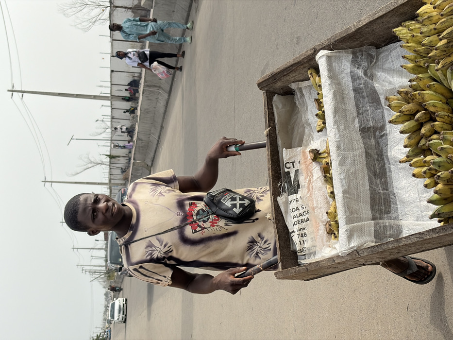 The image captures an outdoor scene, likely a street or roadside market, under bright, possibly hazy, daylight. The main subject is a young man with dark skin and short, dark hair, positioned in the foreground, looking directly at the camera with a neutral expression. He is wearing a light-colored, short-sleeved t-shirt with a prominent dark-toned graphic or pattern, and a black shoulder bag with a white arrow logo (resembling the "OFF WHITE" brand) slung across his chest. He is holding two dark-colored handles or poles, indicating he is pushing or standing with a cart. His bare arms and dark-colored flip-flops are visible.
To the man's right (viewer's left), is a rustic wooden pushcart. It is heavily laden with numerous bunches of yellow, ripe bananas, which are visibly stacked both on top and within the cart. Some of the bananas inside the cart are partially covered by white, woven plastic sacks.
In the background, there's an asphalt road stretching into the distance. Several people are seen walking along the road, and others are gathered or seated on a lower level beyond a concrete barrier or wall that runs alongside the road. Tall utility poles with power lines punctuate the background against a light, hazy sky. Sparse, bare-branched trees are also visible in the distance. A white car is partially visible further down the road.
The visible text on one of the white plastic sacks in the cart includes: "A", "AZIO", "R", "CT", "ging", "GA S", "ALAGE", "IGERIA", "Line:", "748", "11", and "CS@".