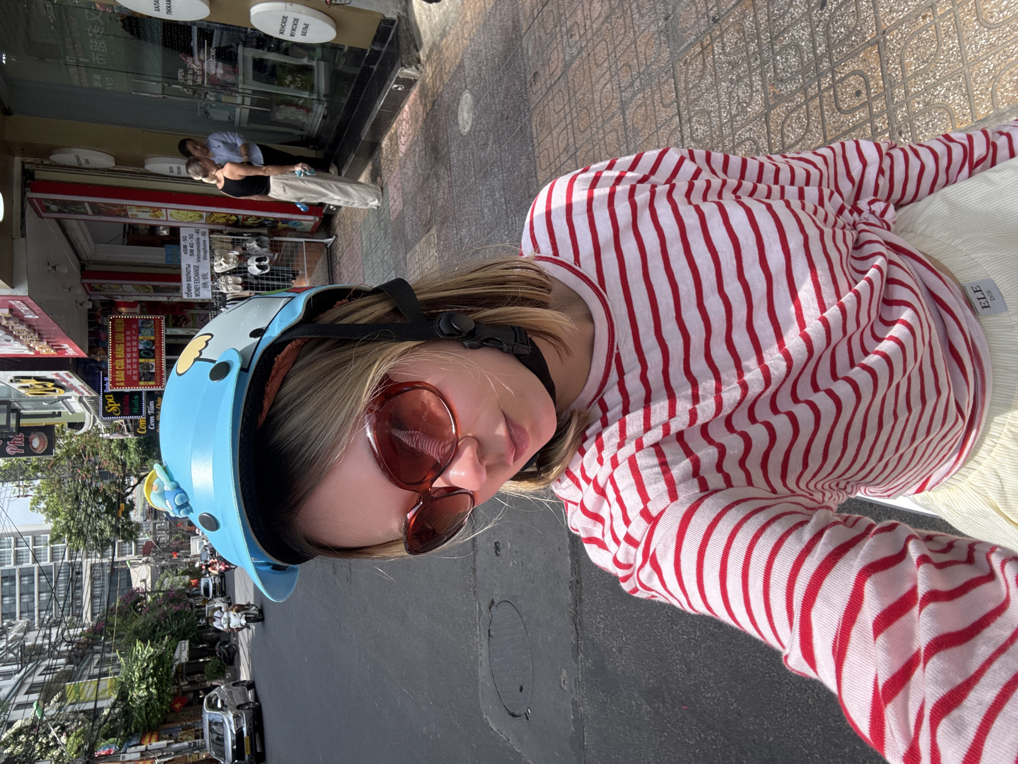 A young woman wearing a blue helmet and sunglasses is taking a selfie on the streets of Nha Trang Ward, Vietnam. She is wearing a red and white striped shirt. The street behind her is busy with traffic, including scooters and cars. Shops and signs line the sidewalk on either side of the street. The weather appears to be sunny. The signage includes texts written in multiple languages, including Vietnamese, Chinese and Russian.