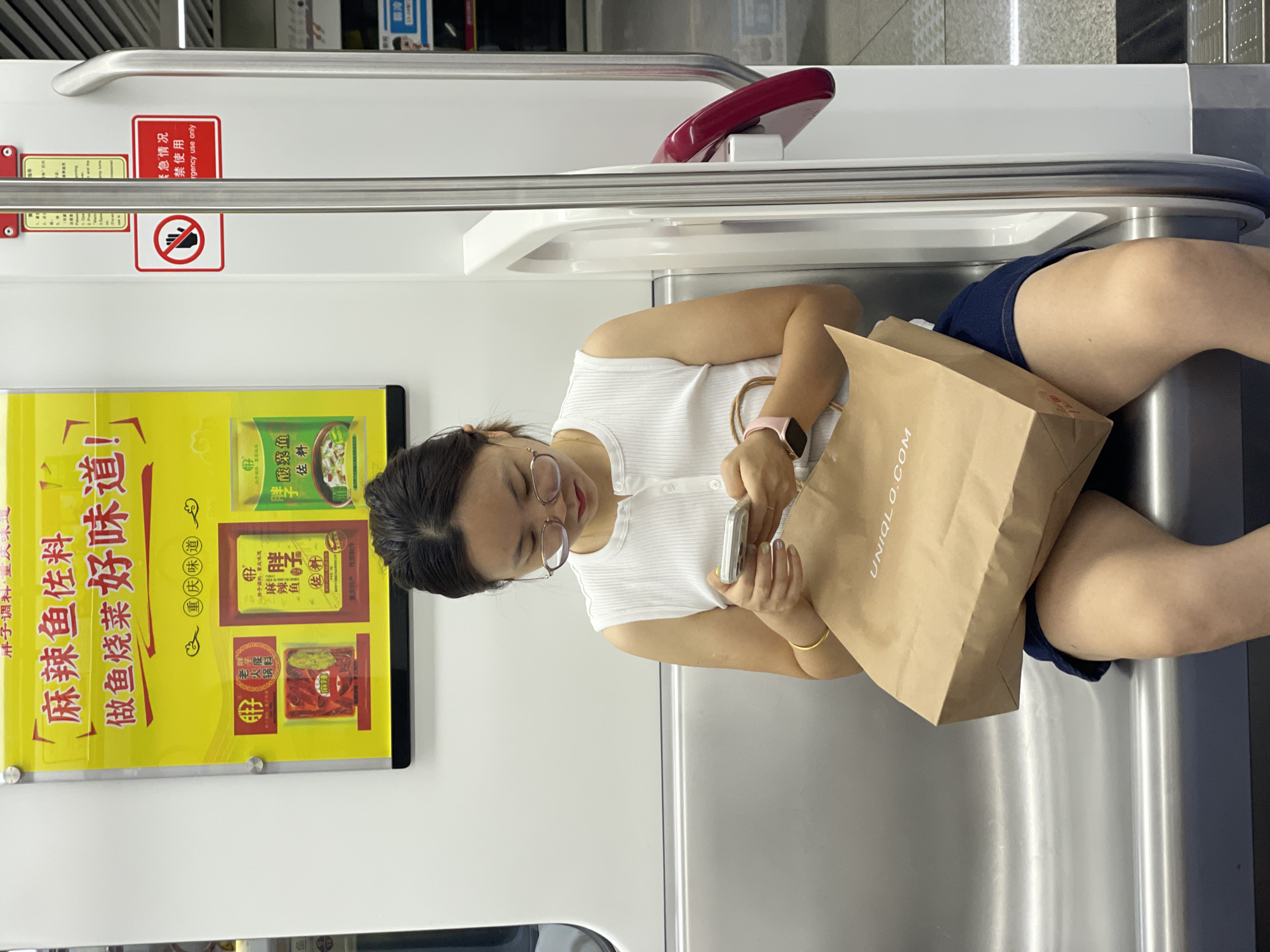 A woman with glasses is sitting on a metal bench in what appears to be a subway car, looking at her smartphone. She is wearing a white sleeveless top and a dark blue skirt. She is holding a brown paper shopping bag from UNIQLO. To her left, there is a yellow advertisement poster with Chinese characters. To her right, there is a vertical metal pole and a red handle. The environment appears to be clean and modern.