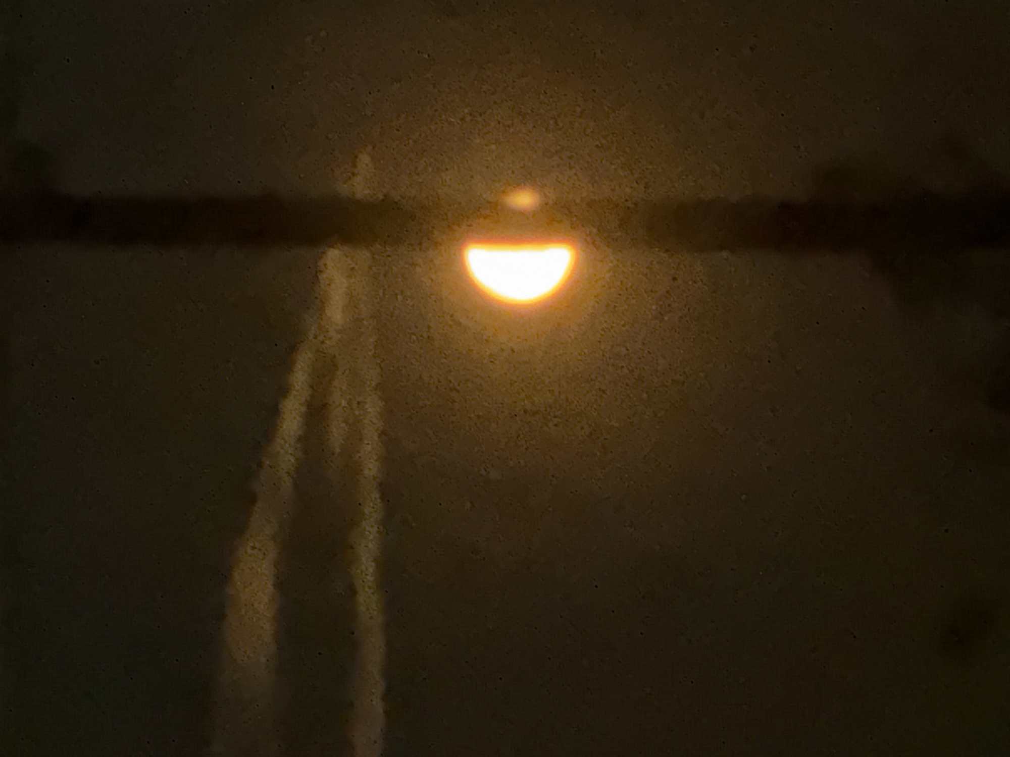 In the city of Baryshevo, Russia, a celestial event is captured. A half-illuminated moon, glowing with an intense orange hue, is visible against a dark, grainy sky. Wispy, pale clouds streak across the upper left portion of the frame. A dark, vertical structure, possibly a tower or antenna, bisects the image, partially obscuring the moon. A smaller, fainter celestial body or reflection is positioned to the right of the moon. The overall scene is dimly lit, with the primary light source being the moon itself.
