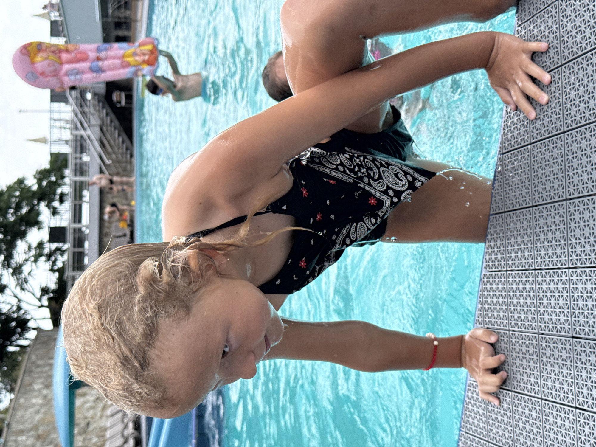 In the city of Massandrivskyi village council, Ukraine, a young blonde girl in a black bandana-print swimsuit is in a swimming pool. She is leaning forward, partially submerged in the water, with her hands gripping the textured grey edge of the pool. Water droplets are visible on her skin and hair. Her face is turned downwards, and her expression appears contemplative. In the background, the blue water of the pool is disturbed by ripples. Other swimmers are present in the pool, including a boy holding a pink inflatable toy and several adults further back. The pool area features outdoor seating and a staircase leading to a higher level. The sky is overcast.