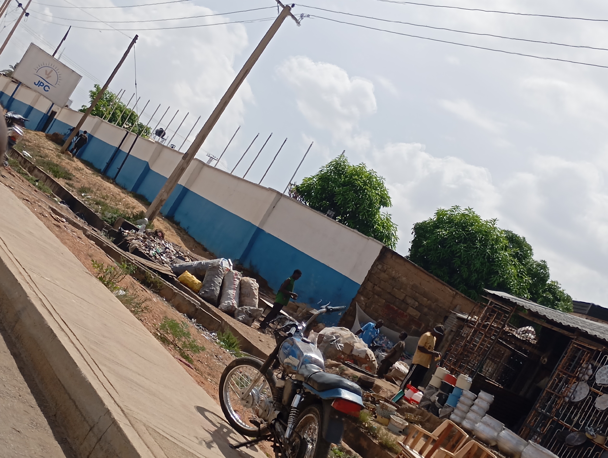 The image captures a vibrant street scene, likely in an urban or semi-urban area of Jos, Nigeria, under a bright, cloudy sky, indicating daytime. The perspective is slightly angled, looking up a gentle incline.
On the left, a concrete sidewalk borders a dirt path, running parallel to a long wall painted white with a prominent blue stripe along its base. This wall is topped with anti-climb spikes. A white rectangular sign is mounted on the wall, featuring the blue letters "JPC" and a sunburst-like logo. A person in a dark shirt is seen walking past this sign.
Further along the dirt path, numerous large, grey and white sacks, seemingly filled with various materials (possibly recyclables), are piled up. A person in a green shirt stands among these sacks, and another in a blue shirt is partially visible behind some materials.
In the foreground, a blue and silver motorcycle is parked or stopped, facing towards the right side of the frame. Its details, including spoked wheels, are clearly visible.
To the right, a makeshift roadside stall or shop, characterized by a corrugated iron roof and metal grilles, displays various household items such as pots, pans, and plastic containers. A person in a yellow shirt stands near this stall, appearing to attend to the goods, with another individual in a brown top partially visible nearby.
Throughout the scene, scattered piles of debris, discarded items, and additional sacks suggest activities related to informal recycling or storage. Green trees with dense foliage are visible above the walls and structures. Overhead, several wooden utility poles carry multiple power lines across the sky. The overall image depicts a lively daily scene with informal commerce and everyday activities.