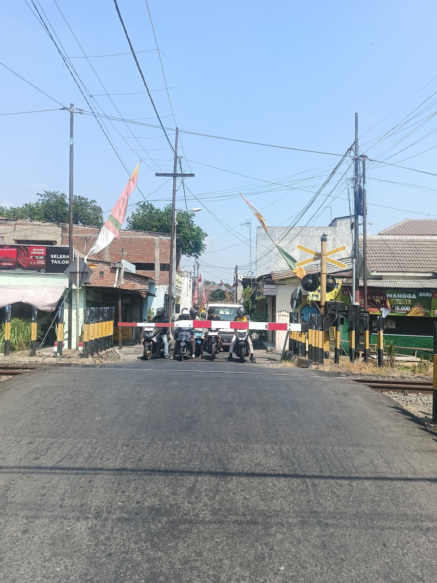 The image shows a railroad crossing in Gedangan, Indonesia. A red and white barrier is lowered, indicating that a train is approaching or passing. Several motorbikes are stopped at the barrier, waiting to cross. In the background, there are buildings, utility poles with many wires, and trees. The sky is clear and blue. The road surface is asphalt. There are yellow and black striped poles on either side of the crossing. A railroad crossing sign with diagonal lines is visible.