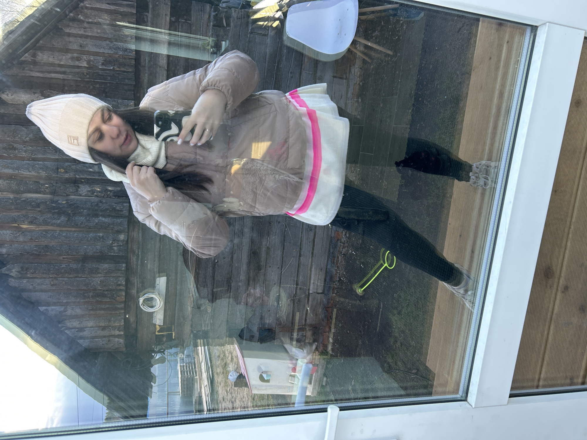 The image captures a selfie reflection of a young woman, taken through a window or glass door. The woman, positioned centrally, is holding a smartphone with a black and white patterned case, presumably taking her photo. She has long, dark hair and is dressed in a light-colored (cream or beige) puffer jacket, a matching knitted beanie adorned with a small, stylized "TL" logo, and a coordinating turtleneck or neck warmer. She also wears dark leggings and light-colored sneakers. A silver ring is visible on her left-hand ring finger.
Behind the reflection of the woman, an outdoor scene is visible through the glass. This includes a rustic wooden building with horizontal planks, possibly a shed or an old structure. The ground features a mix of grass and dirt, where a vibrant yellow-green gardening tool, resembling a trowel or small rake, lies. A small, multi-colored birdhouse with white, pink, and blue details is also on the ground. Faintly visible in the reflection of the interior space is a white plastic chair or stool.
The scene takes place during the daytime, with natural light illuminating the environment. The woman's warm attire suggests cool or cold weather conditions. The image combines the reflected indoor space with the transparent view of the outdoors, creating a layered visual effect. The subject is standing upright. The location context provided is Chaikovsky, Russia.