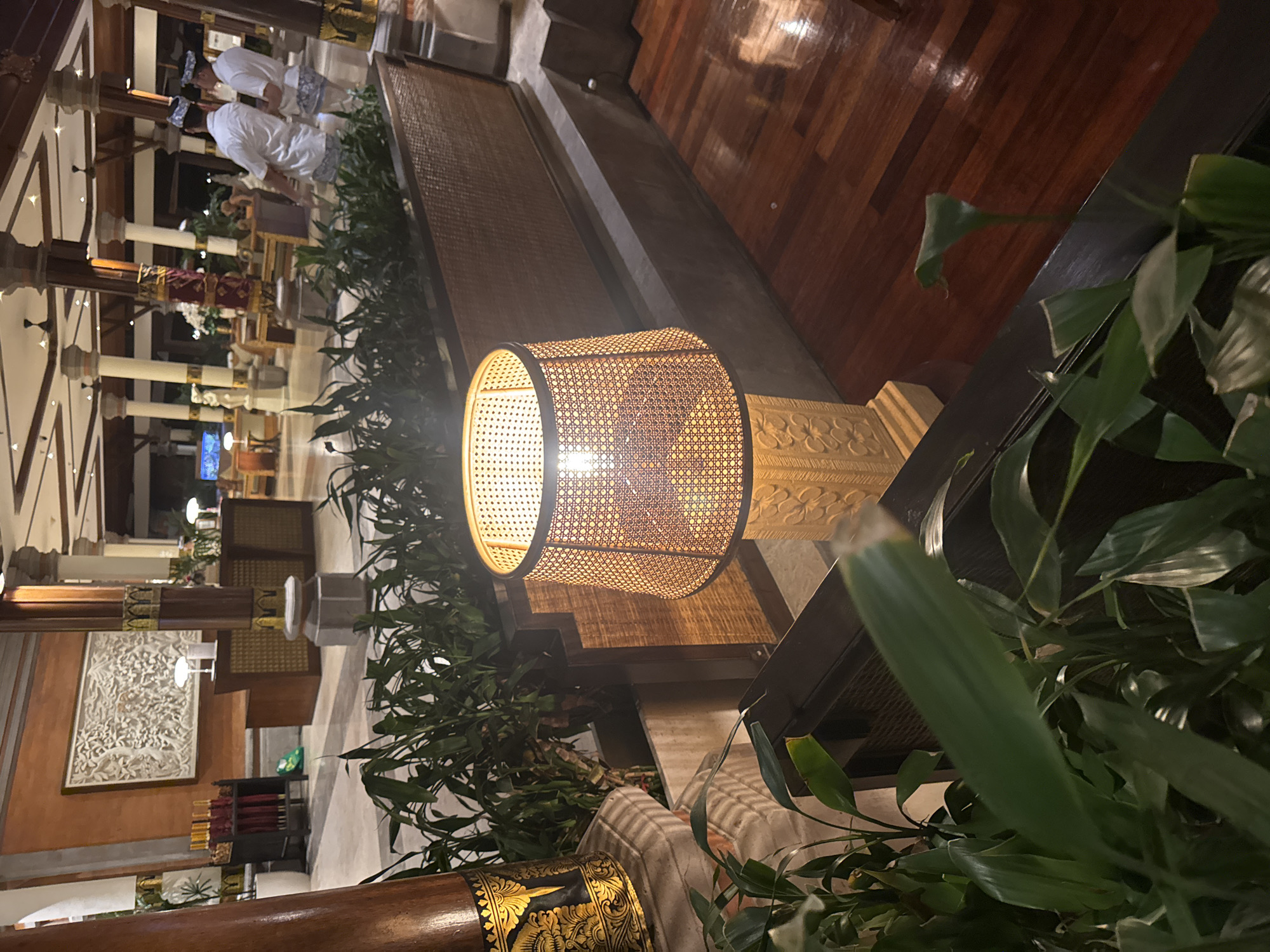 The image captures the interior of what appears to be a hotel lobby in Kuta Selatan, Indonesia. A prominent feature is a tall floor lamp with a woven lampshade emitting a warm glow, positioned in the foreground. To the left of the lamp, lush green plants are visible, adding to the ambiance. In the background, a reception desk area is seen with decorative elements and what looks like an artistic wall hanging. Two individuals dressed in traditional attire, one in a white shirt and the other in a white tunic and sarong, are standing near the reception area, possibly staff members. The lobby has a high ceiling with decorative columns and ambient lighting, suggesting a luxurious and traditional setting. The flooring is a dark, polished wood.