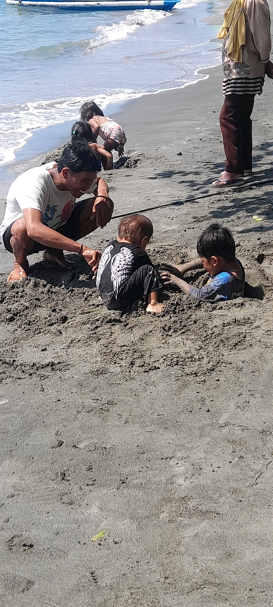 In Pasir Putih, Indonesia, a sandy beach scene unfolds under bright sunlight. A man in a white t-shirt and dark shorts kneels on the sand, facing two young children playing. One child, wearing a black shirt with white wing designs and black pants, is seated in the sand. To their right, another child, in a blue long-sleeved shirt, is partially buried in the sand up to their chest, actively playing. In the background, closer to the shoreline, a woman in a light-colored top and dark pants stands, observing the scene. Further back, near the water's edge, two younger children are also engaged in play on the wet sand. The ocean waves gently roll onto the beach, and a blue and white boat is visible on the water in the distance. The overall environment is one of a casual, family-oriented beach day.