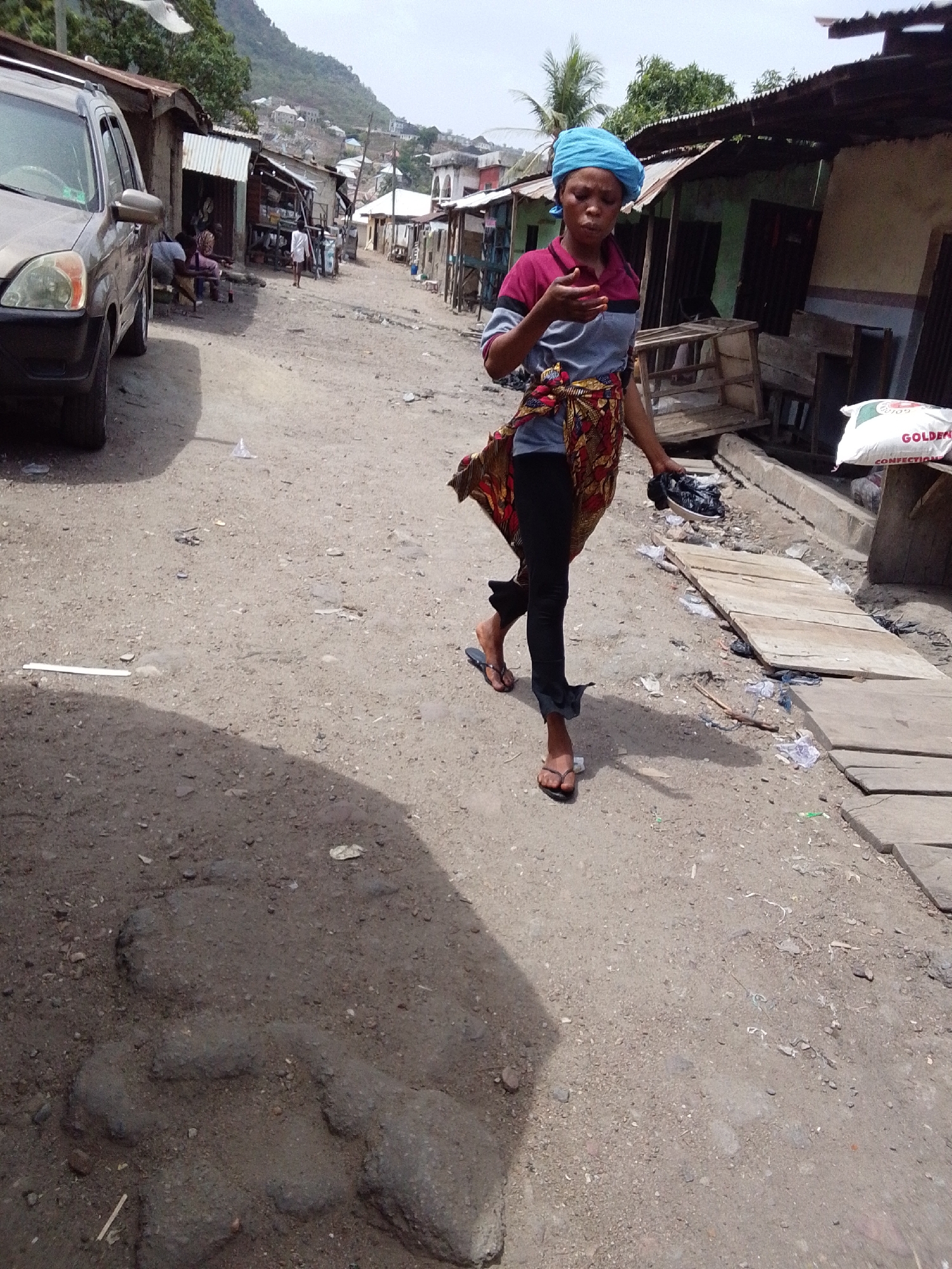 A woman is walking in the street of Lokoja, Nigeria. The woman is wearing a blue headwrap, a maroon and gray shirt, black pants, and flip-flops. She has a patterned fabric tied around her waist and is carrying a pair of black shoes in her right hand. The street is unpaved. There are buildings on both sides of the street, some are shacks. A light-colored SUV is parked on the left side of the street. There are a few people in the background. A hill is visible in the distance. A bag of confectionary is to the right of the woman.