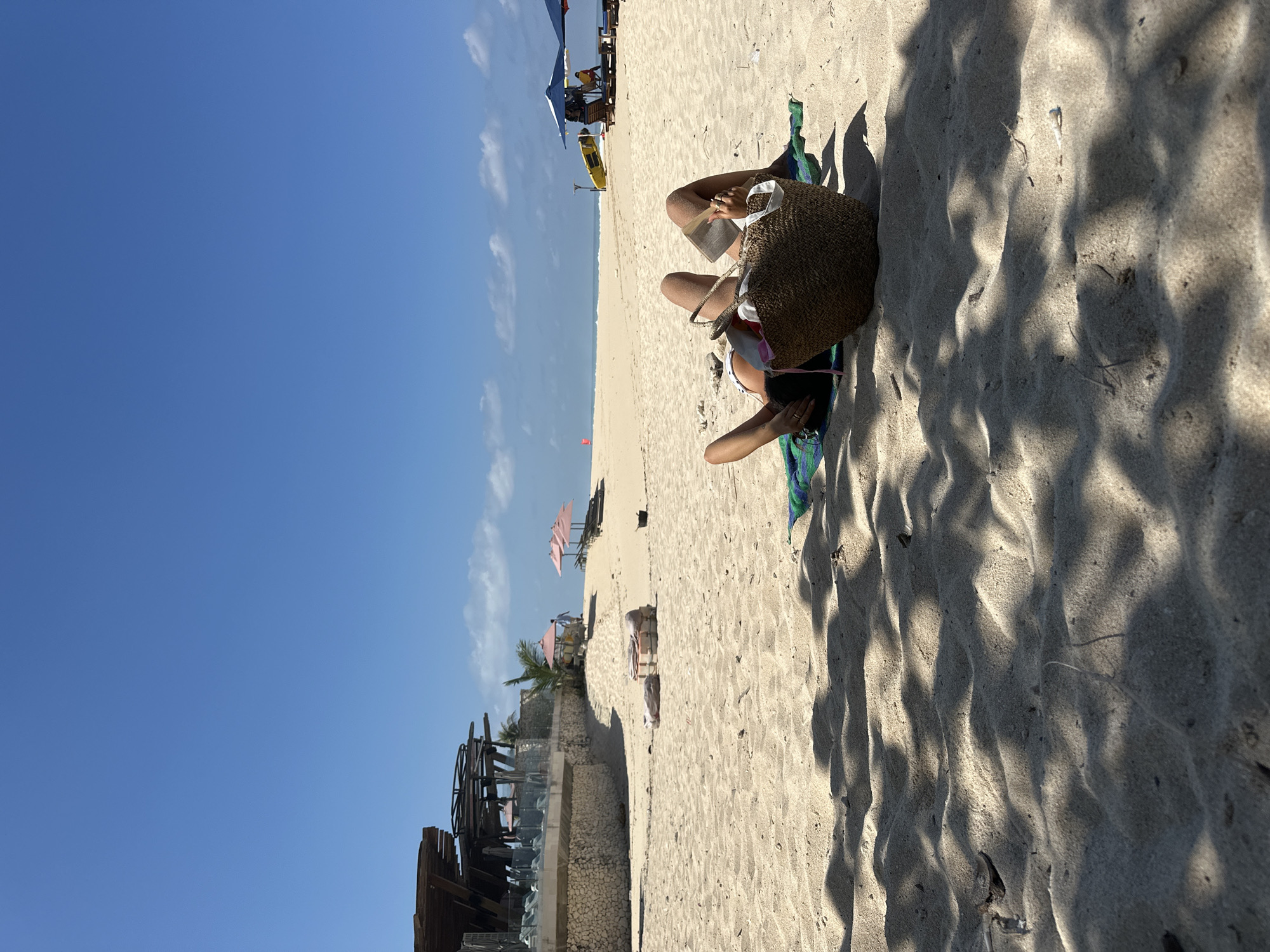This image captures a serene beach scene, likely in Kuta Selatan, Indonesia, on a bright, sunny day. The main subject is a person relaxing on light-colored sand, lying on their stomach or side, facing away from the viewer. They are wearing a light swimsuit top and appear to be reading a book or looking at a device held in their hands. A large, woven straw beach bag rests beside them, and a striped beach towel (blue, green, and white) is visible underneath.
The setting is a picturesque beach with a vast expanse of light, fine sand. In the mid-ground, a narrow strip of the blue ocean is visible, leading to the horizon. Further back on the beach, several beach umbrellas (some blue, some pink) and chairs are set up, suggesting a resort or public beach area. A yellow kayak or small boat is also present near the water's edge. To the far left, a multi-tiered structure made of wood and light-colored stone or concrete is visible, hinting at a beach club or hotel.
The clear blue sky, with just a few wispy white clouds, indicates good weather. Strong sunlight casts prominent, intricate shadows across the sand in the foreground and mid-ground, suggesting it is mid-day. The overall atmosphere is one of relaxation and leisure. No visible text can be discerned in the image.