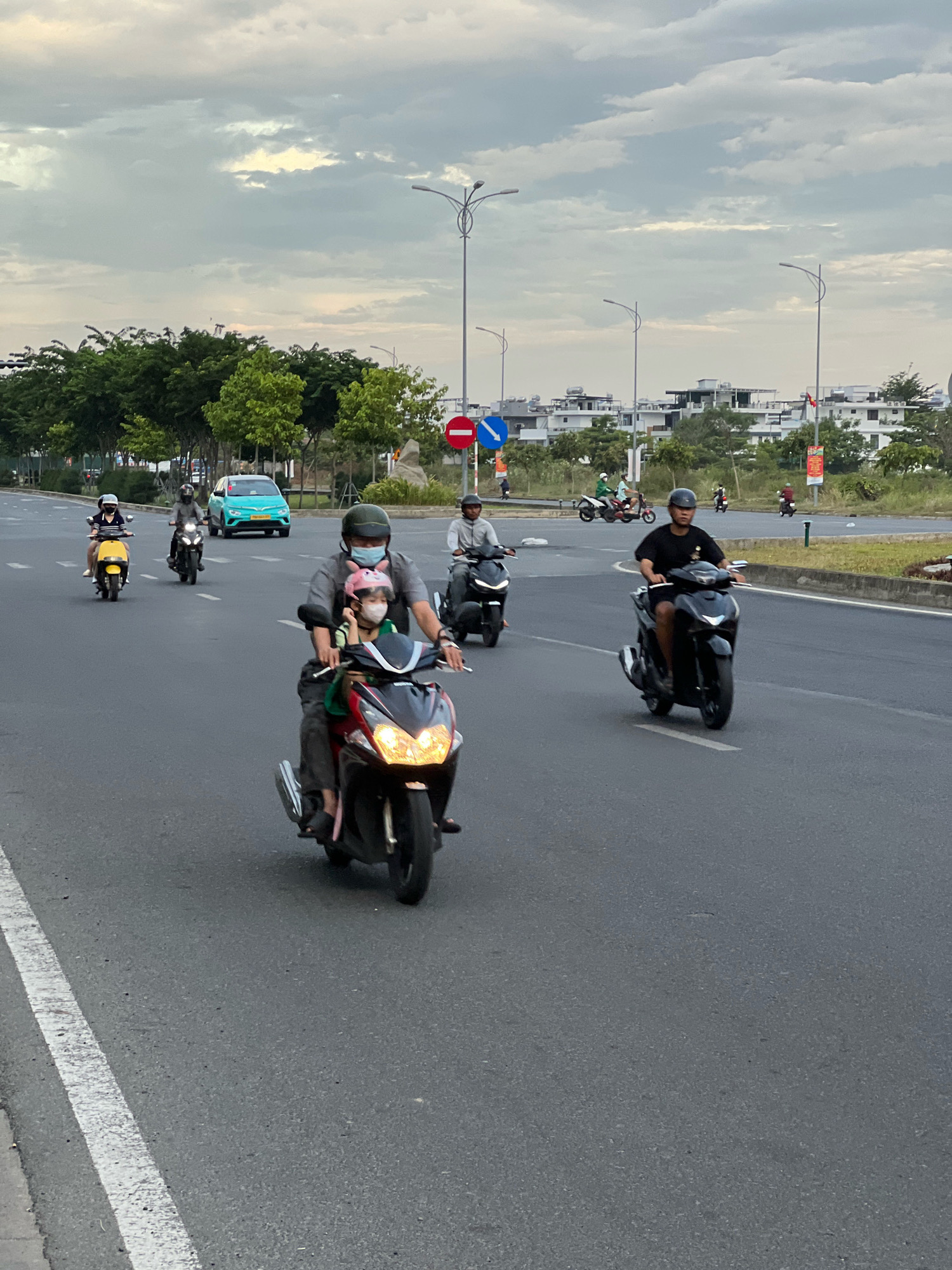 The image captures a scene on a multilane road in Nha Trang, Vietnam, likely during overcast daylight. Several individuals are operating and riding on motorcycles and scooters. In the foreground, a man wearing a face mask and a dark gray shirt, along with a young child in a pink helmet and face mask, are riding a red and black scooter. To their left, a woman in a striped shirt rides a yellow scooter, and behind her, another person rides a dark scooter. Further back on the road, a bright turquoise car is visible, along with other motorcycles and scooters. The background features a line of trees and a modern urban landscape with white buildings and streetlights. The road surface appears to be asphalt, marked with white lane lines.