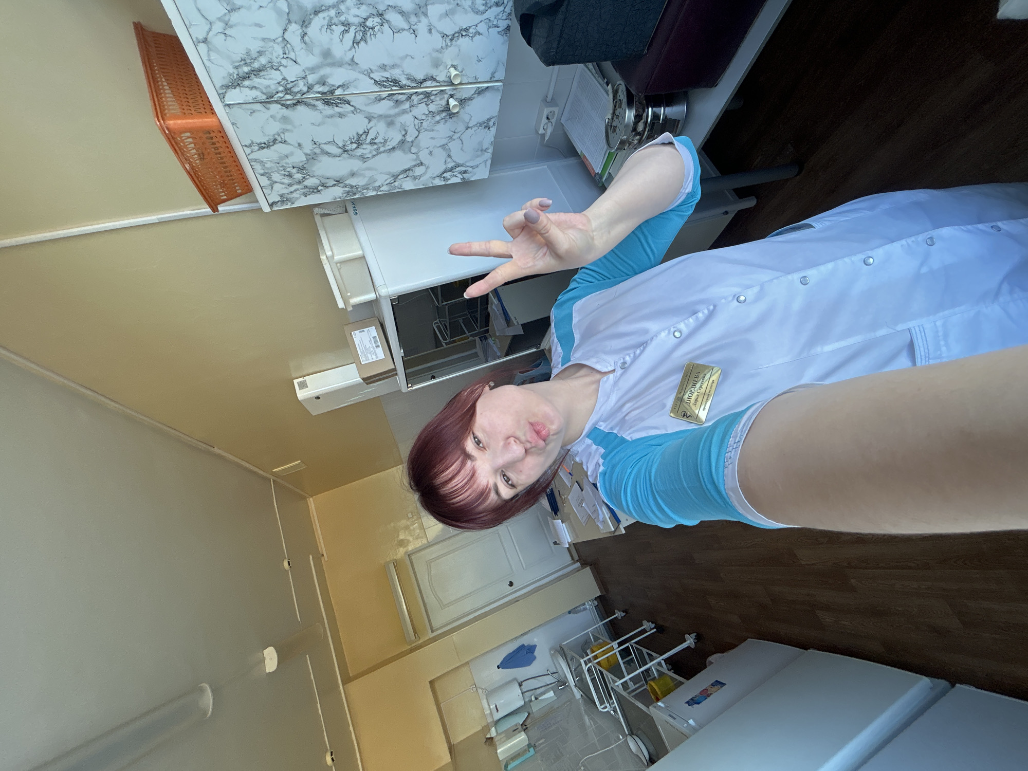 A selfie shows a woman in a white and light blue uniform with a name tag. She has short auburn hair with bangs and is making a peace sign with her hand. She appears to be in a medical or dental office. To the right of the person are two beds and to the left are white cabinets, a storage basket, and various pieces of equipment. Based on the name tag in Cyrillic, it is likely taken in the city of Фоки, Russia.