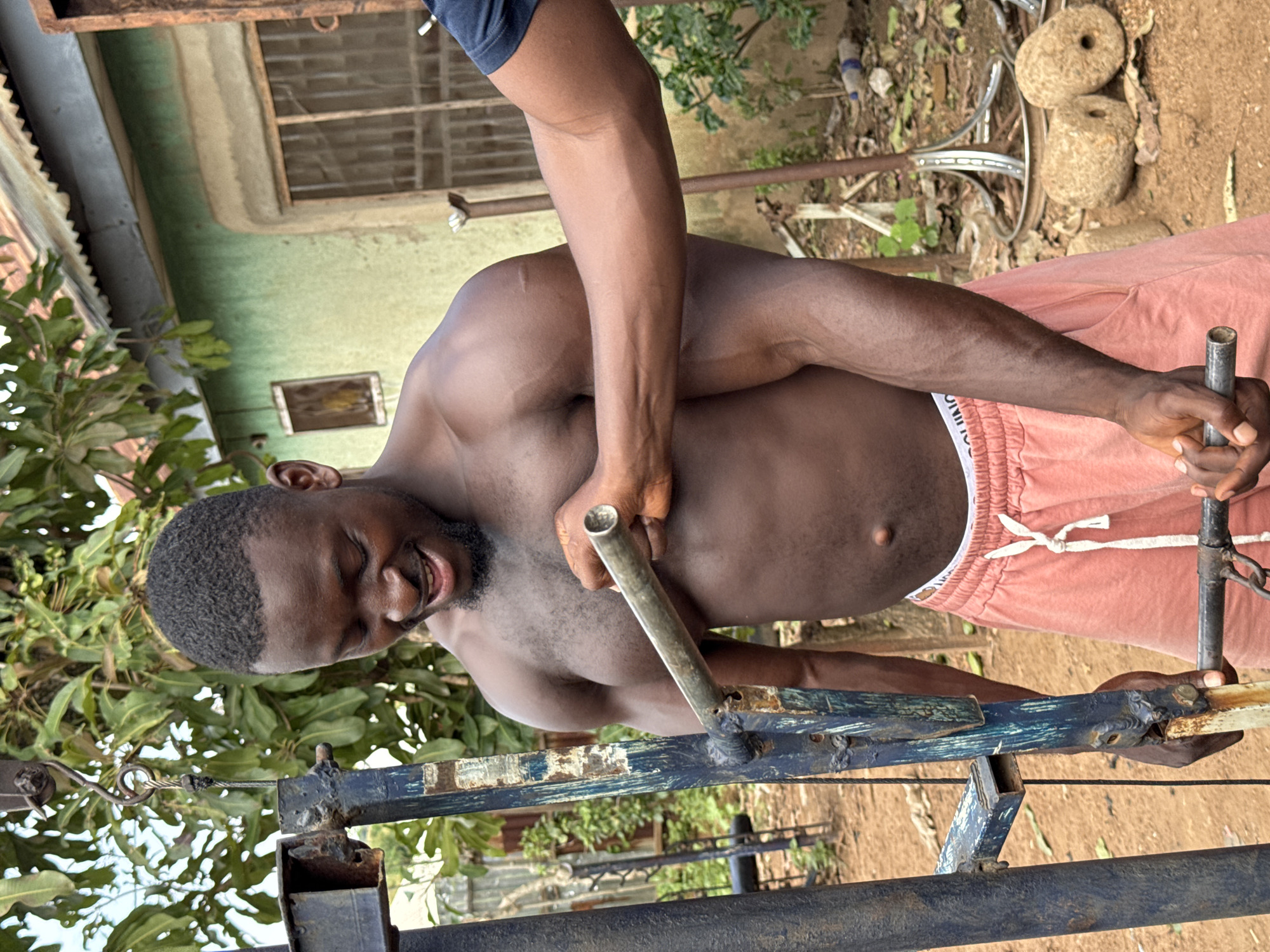In a somewhat rough and utilitarian outdoor setting, likely in Akwanga, Nigeria, a muscular Black man is engaged in a physical activity. He is shirtless, wearing pink athletic shorts with white trim. His body is turned to the side, and he appears to be pulling or lifting a metal bar or tube with his left hand. His right arm is bent, and his hand is positioned higher up on the same bar. His head is tilted back, eyes closed, and he has a strained expression, suggesting exertion. He has a short haircut and a beard.
The background reveals a light green painted wall, possibly of a building, with a dark-framed window. To the left, there are green leafy plants. In the upper right portion of the image, two large, rough-hewn stones with circular holes are visible, hinting at their potential use as weights or part of some apparatus. The ground appears to be dirt. The overall scene has a raw, unpolished feel, suggesting an informal training space. The lighting indicates it is daytime, likely under natural sunlight, although the exact time of day is difficult to ascertain. No visible text is present in the image.